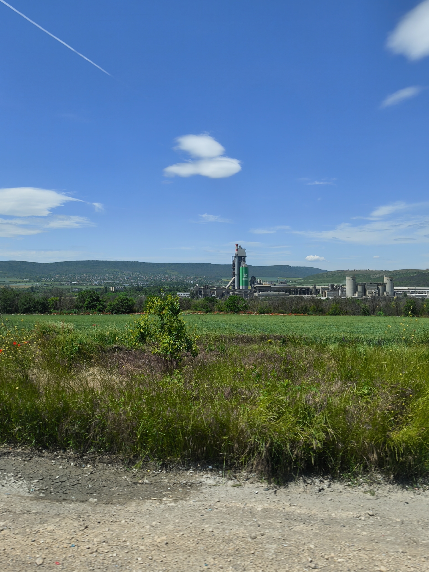 The image presents a landscape in an unidentified location, featuring a clear blue sky with a contrail from an airplane and scattered clouds. A large industrial complex, possibly a factory or plant, dominates the midground. This complex is situated amidst a field of green vegetation and sparse trees. The background includes rolling hills and a town or residential area nestled in the distance. In the foreground, there's a rough, dirt road bordering a patch of unkempt, overgrown grass.