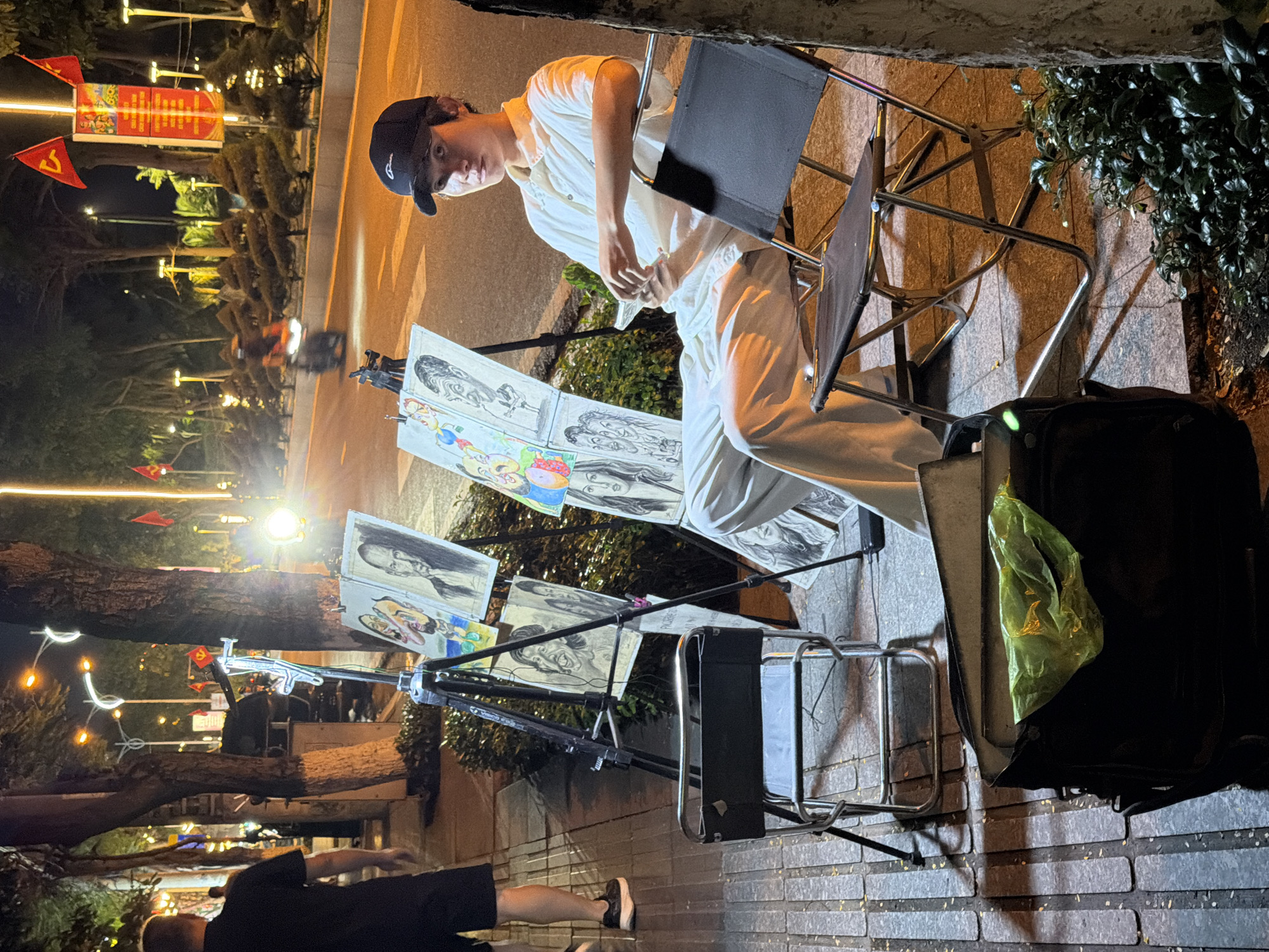 The image depicts an outdoor scene at night in Nha Trang, Vietnam, possibly on a street or promenade. In the foreground, a young man wearing a white polo shirt and a black baseball cap is seated on a folding chair, holding a smartphone. He is looking towards the camera with a neutral expression. In front of him, on easels, are several charcoal sketches of faces, some of which appear to be caricatures. To his left, another empty folding chair is positioned, and a black bag with a bright green plastic bag inside rests on the ground. The background shows a street with traffic, including motorcycles, and illuminated trees and streetlights. Red flags and banners are visible, suggesting a public or festive area. The ground appears to be paved with rectangular stones, wet from rain or cleaning. The overall atmosphere is that of a nighttime street scene with an artist and his work displayed.