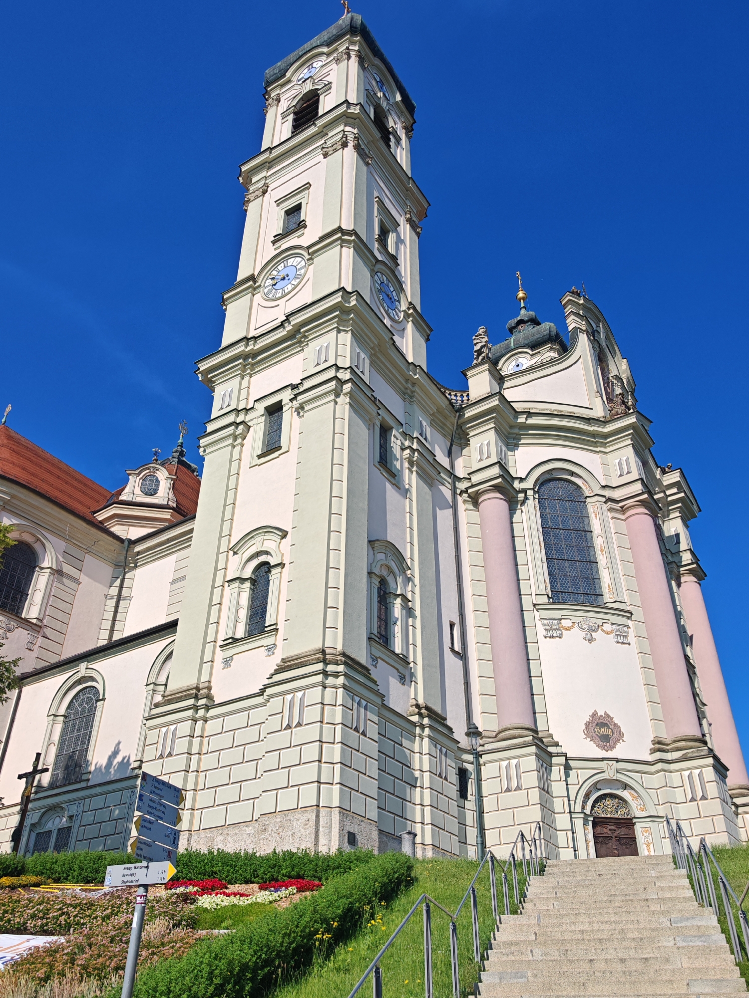 The image shows the Basilica of St. Alexander and St. Theodore in Ottobeuren, Germany, under a clear blue sky. The focus is on the imposing facade and bell tower of the basilica, constructed with a pale yellow and white stucco. The bell tower is tall and ornate, featuring multiple clocks with blue numerals and Roman hour markers. Large windows with decorative stained glass are visible on various levels of the building. The lower part of the basilica has a series of arched windows and a prominent entrance at the top of a wide stone staircase. The staircase is flanked by metal railings and a grassy slope adorned with colorful flowerbeds in various shades of red, pink, white, and yellow. A cross stands to the left of the stairs, partially obscured by vegetation. A directional signpost with multiple yellow and blue arrows is positioned on the slope, indicating various destinations and walking times. The architecture is baroque in style, characterized by its grandeur and elaborate ornamentation. The scene appears serene and well-maintained, suggesting a place of worship and tourist attraction.
