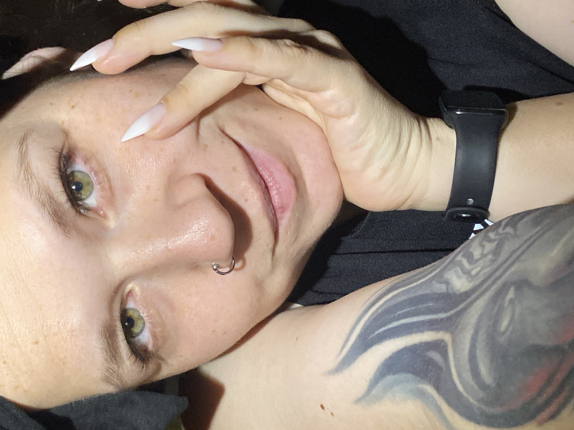A close-up, angled portrait shows a fair-skinned woman with freckles and green eyes. She has a silver hoop nose piercing and white, pointed manicure. Her hand is partially covering her face. On her wrist is a black smartwatch. A portion of a dark-colored tattoo is visible on her upper arm/shoulder. She is wearing a dark shirt. The scene is indoors. There is no indication of location in the city of Chaikovsky, Russia.