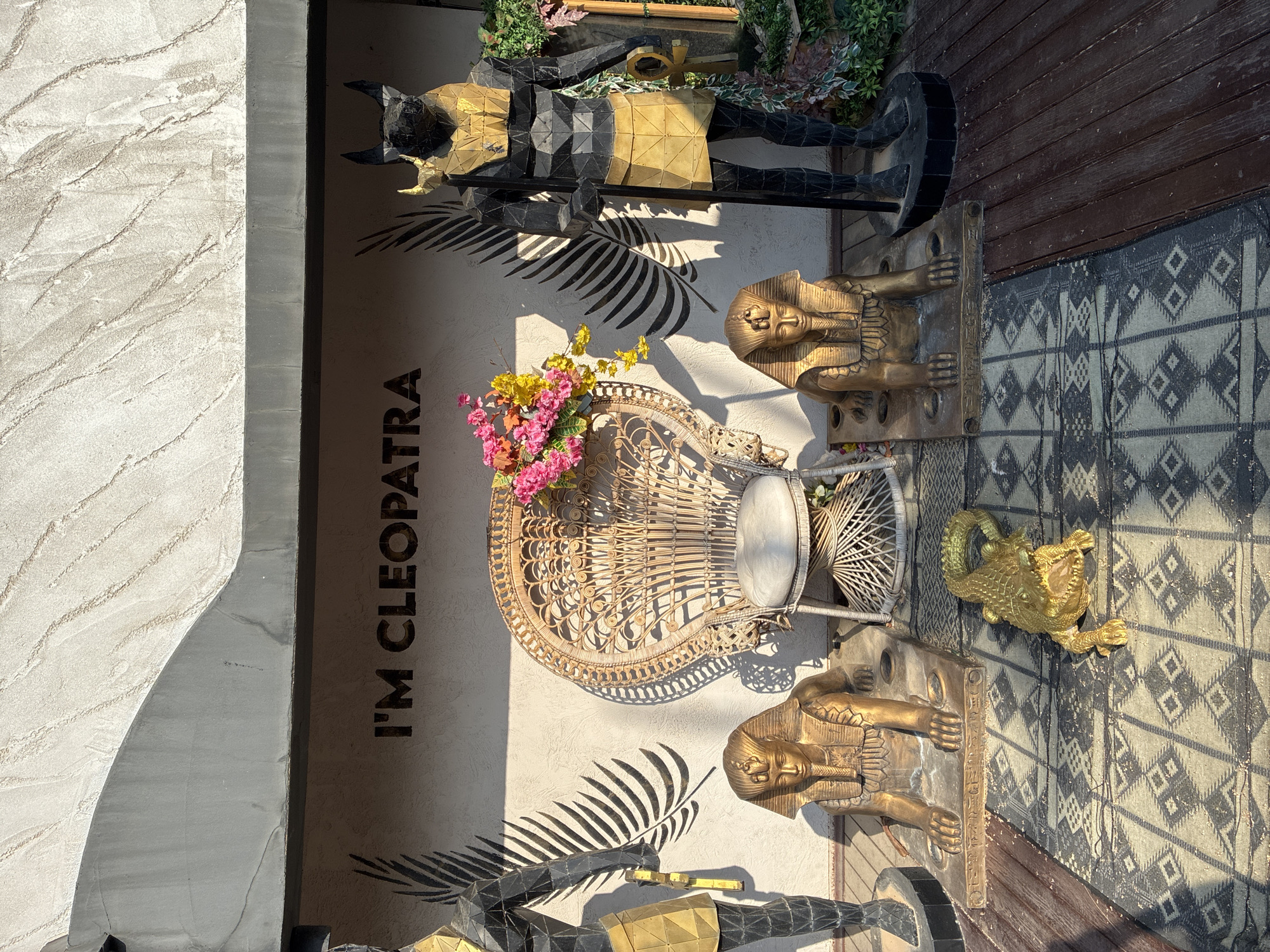 The image shows an outdoor patio decorated with an Egyptian theme, likely part of a business or restaurant. The word "I'M CLEOPATRA" is painted in large, dark letters on a textured beige wall in the background. To the left and right of a prominent wicker peacock chair are two golden sphinx statues. Behind the chair, a bouquet of pink and yellow flowers is displayed. Further to the right stands a tall statue of a figure with the head of an animal, possibly Anubis, holding a staff. In front of the sphinx statues, on a patterned rug, lies a golden alligator statue. The patio floor is made of dark wood planks. The scene is illuminated by bright sunlight, casting distinct shadows. This scene is located in the city of Alanya, Turkey, identified by the style and decor.