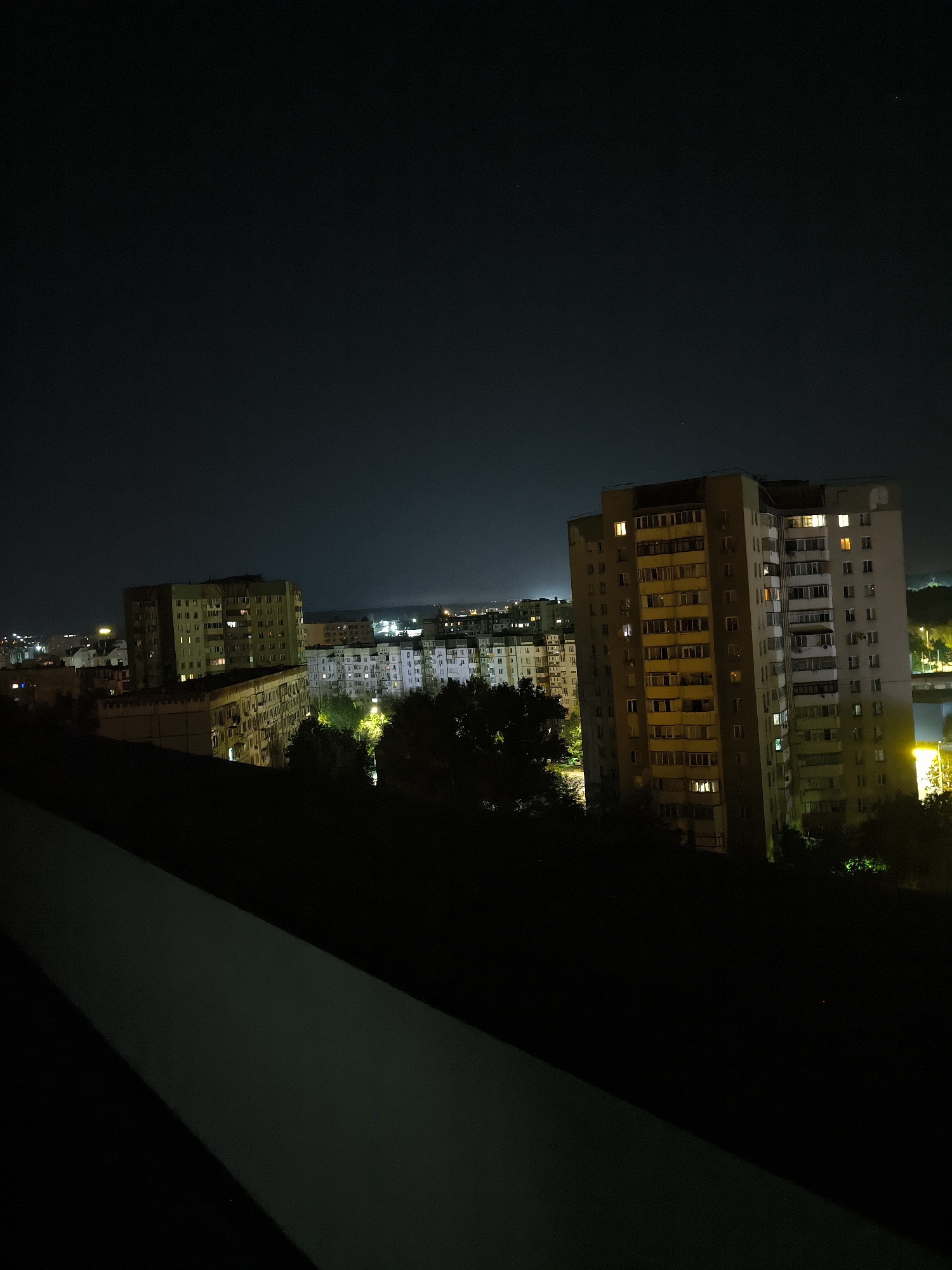 This is a night-time, medium-shot, eye-level photograph of a residential area in the city of Chișinău, Moldova. The foreground is dominated by a dark, angled surface, likely a balcony railing or a ledge, which casts a shadow across the bottom left of the frame.
In the midground, several multi-story apartment buildings are visible. The most prominent is a tall, rectangular building on the right, with numerous windows illuminated from within, suggesting habitation. Its facade is a light brown and grey color. To its left, another, slightly shorter apartment building stands, also with lit windows and a yellowish-green hue. Further to the left and in the background, more apartment buildings can be seen, some appearing lighter in color and with fewer visible lights.
Trees and foliage are scattered throughout the midground and foreground, silhouetted against the lit windows and the dark sky. Some trees appear taller and denser, while others are more sparse. Streetlights and other urban lighting are visible in the distance, creating a hazy glow on the horizon. The sky is dark and clear, with a few faint stars visible. The overall impression is of a typical urban night scene, with the lights of the buildings creating a sense of life within the darkness.