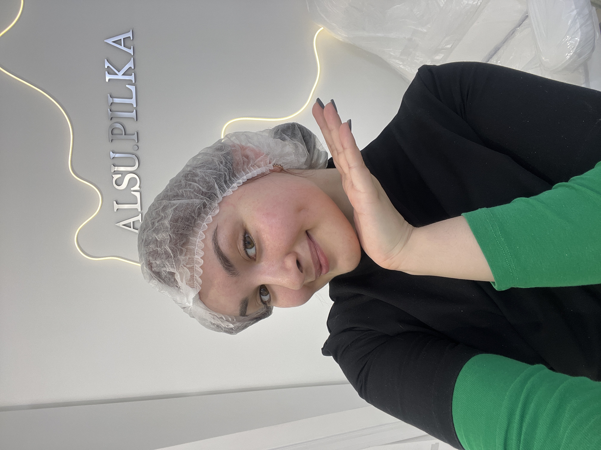 A woman with a hair net and long dark nails poses indoors at "ALSU.PILKA" in Neftekamsk, Russia. She wears a black top with green sleeves. Light outlines the name on the wall.