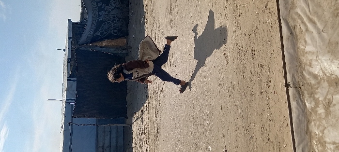 A single child is depicted running from left to right across a dirt path in Khan Yunis, Palestinian Territory. The child, with dark, curly hair, wears a brown jacket over a lighter garment that shows the partial word "SELF," dark trousers, and open-toed sandals. A light-colored blanket or fabric is draped over the child's left shoulder and back. Strong sunlight from the left casts a distinct shadow of the child on the ground to their right. In the background, several makeshift structures are visible under a clear blue sky with sparse clouds. These structures appear to be constructed from various materials, including light-colored tarpaulin or sheeting, corrugated metal, and dark patterned fabric. Upright poles or antennas protrude from the rooftops of some structures. The immediate foreground shows a lighter, textured ground covering, separated from the dirt path by a dark line.