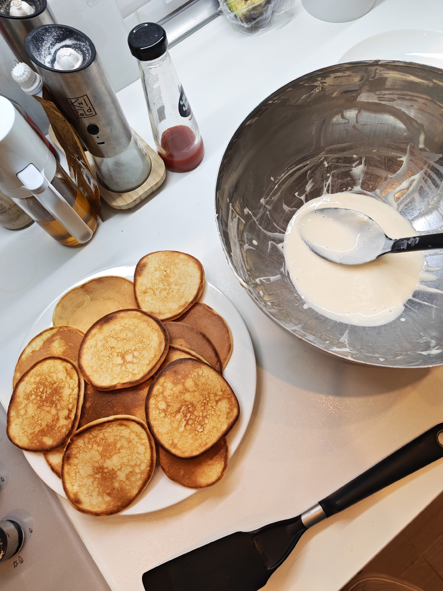 This image shows a close-up, high-angle view of a kitchen counter. In the foreground to the left, a white plate is piled high with golden-brown pancakes. To the right of the plate, a large stainless steel mixing bowl contains a pale, viscous liquid, with a black spoon resting in the bowl. The bowl appears to have some of the liquid splattered on its sides. In the background, various kitchen items are visible, including a stainless steel electric spice grinder, a glass bottle with a black cap containing a reddish liquid, and another bottle with a white pump dispenser. A black spatula lies horizontally in the bottom right corner of the frame. The scene suggests the preparation or aftermath of making pancakes. The location in the city of Bataysk, Russia, is not discernable from the visual evidence in the image.