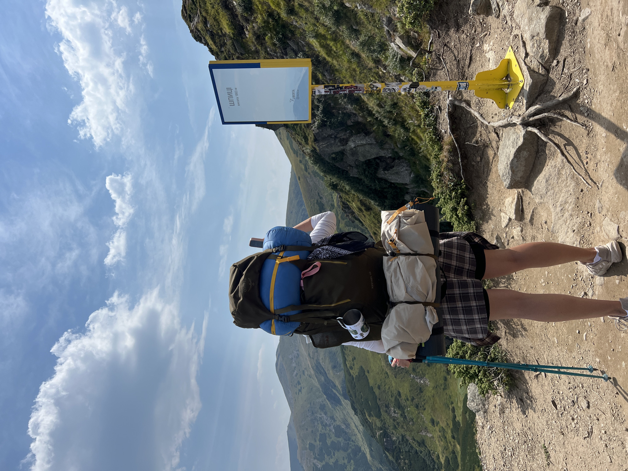 The image shows a person from behind, wearing a large backpack and holding trekking poles. The person is standing on a mountain trail with a scenic view of rolling green hills and mountains under a partly cloudy sky. To the right of the person is a yellow sign with blue and white markings, likely indicating a location or trail information. The sign is decorated with stickers. The foreground shows rocky terrain with some exposed roots. The overall setting is a mountainous landscape, suggesting an outdoor activity like hiking or mountaineering. The sign appears to be in Ukrainian, with text that reads "ШПИЦІ" and "висота 1863 м", which translates to "Shpytsi" and "height 1863 m". Shpytsi is a mountain peak located in the Carpathian Mountains near Vorokhta, Ukraine.