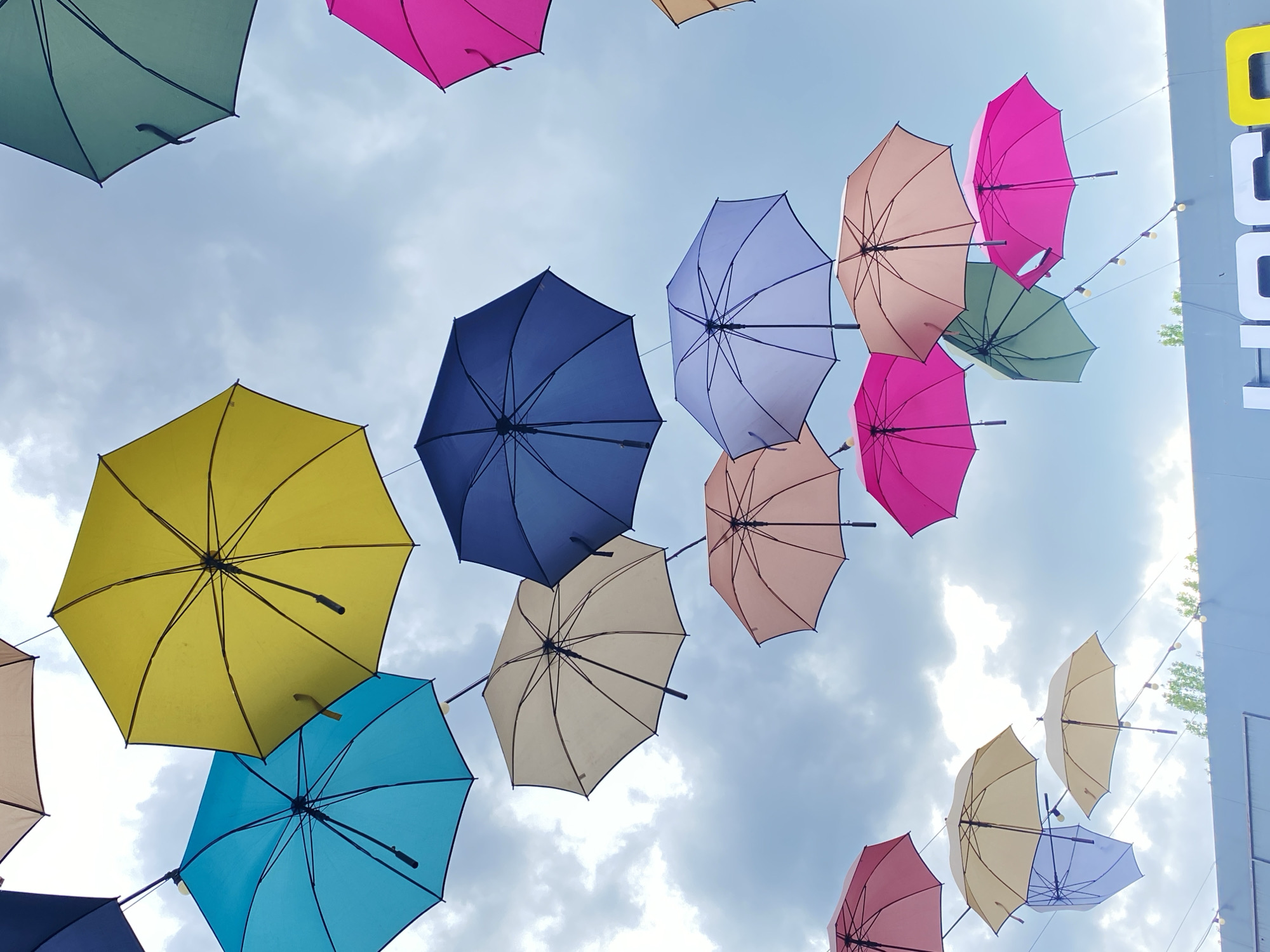 The image shows a view looking up at a sky decorated with numerous colorful umbrellas. The umbrellas, in shades of pink, yellow, blue, green, and beige, are suspended in the air against a backdrop of a cloudy, light blue sky. In the upper right corner, there is a glimpse of a building or structure with white and yellow elements, and some string lights. The location is a city, though the specific location and the identities of any individuals are unknown.