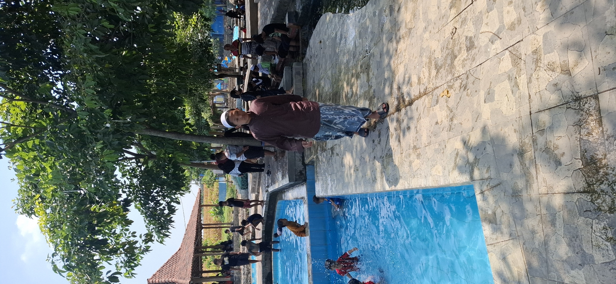 A boy wearing a brown shirt and a white cap, walks on a tiled pathway next to a bright blue swimming pool. Several people are seen in and around the pool, some swimming and others sitting on the edge. In the background, there is a red-tiled roof structure, trees, and a clear blue sky. The scene appears to be a public swimming area on a sunny day.
