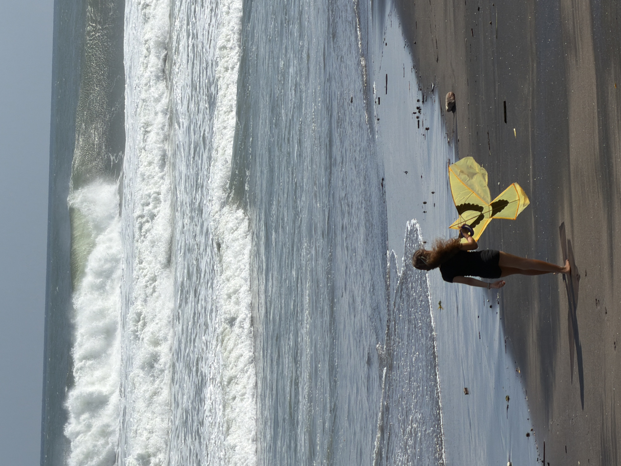 A young woman with long, dark hair walks on a dark sand beach toward the ocean in Mengwi, Indonesia. She is wearing a black short-sleeved top and black shorts. In her right hand, she holds a yellow kite with black markings, shaped like a butterfly. The kite is partially unfurled, suggesting it is about to be flown or has just been flown. The ocean is rough, with large waves crashing near the shore, creating a white foam. The sky above is overcast, with a hazy, gray-blue hue. The sand is dark and appears wet near the water's edge. Scattered debris, possibly driftwood and shells, is visible on the beach. The overall scene depicts a windy day at the beach with a lone figure about to engage in a recreational activity.