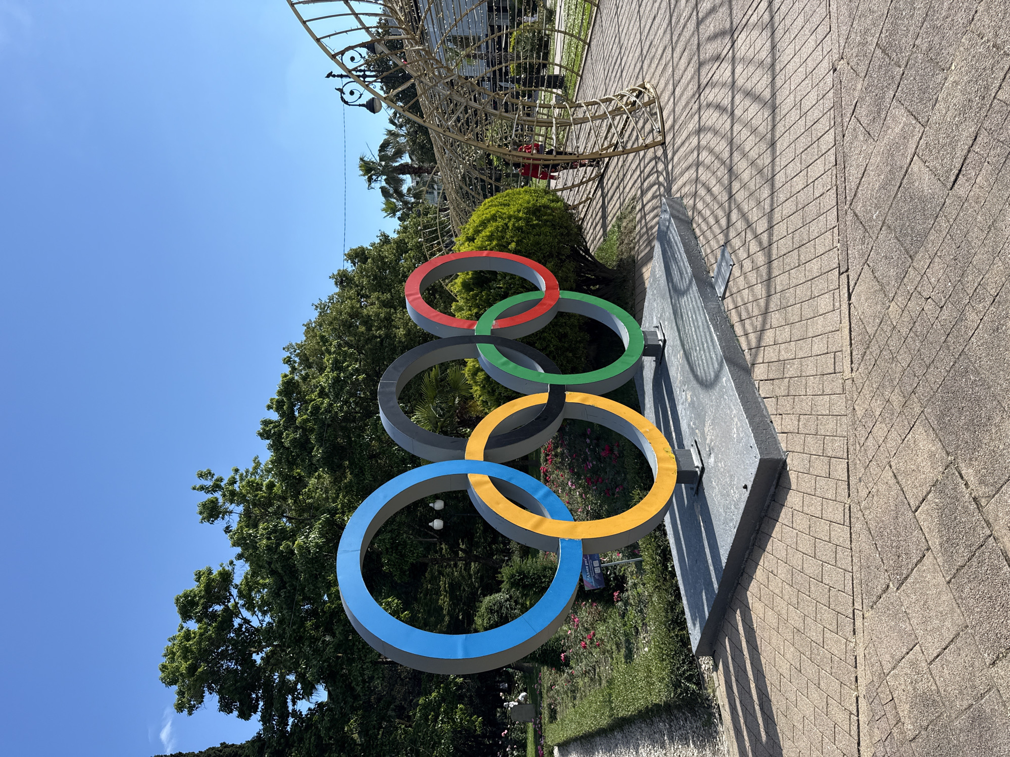 In this sunny outdoor scene in Sochi, Russia, the iconic Olympic rings sculpture stands prominently in the foreground. The sculpture, colored blue, yellow, black, red, and green, is positioned on a gray rectangular base. Behind the sculpture, a lush green lawn is adorned with pink flowers, and mature trees line the background. To the right, a person wearing a red top and patterned pants walks under a large, intricate golden arch structure. The ground is paved with textured tan bricks, and the clear blue sky above indicates pleasant weather.