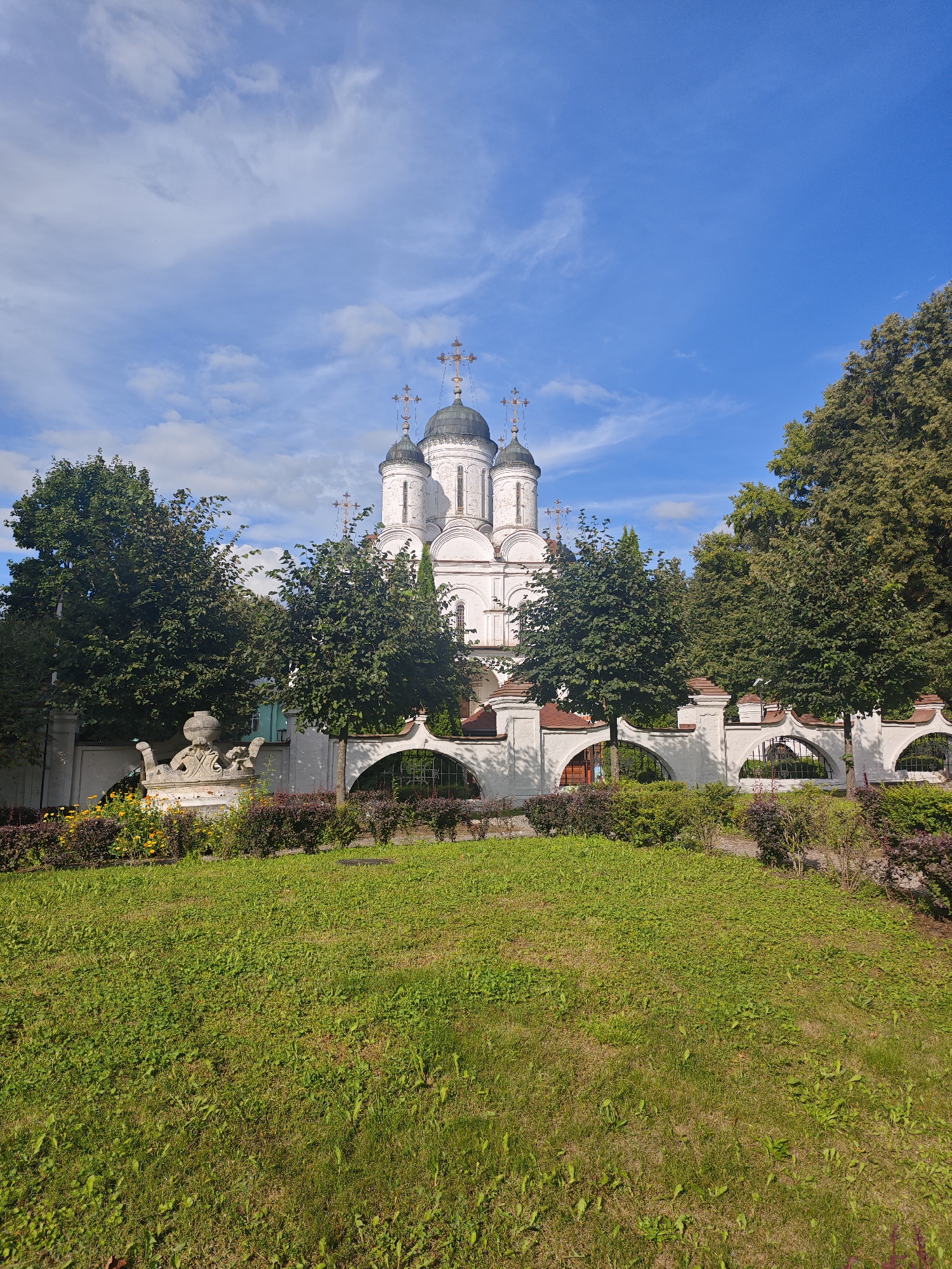 This is a vertical, full shot of the Church of the Resurrection of Christ in Bolshiye Vyazyomy, Russia, under a bright blue sky with scattered white clouds. The church, a white, multi-domed Orthodox Christian structure with gold crosses on its domes, is visible in the background. It is surrounded by a white, ornate fence with arched openings, some of which have decorative metalwork. Mature green trees frame the church and fence, with some foliage extending into the foreground. In front of the fence, a low hedge of dark green and reddish-purple bushes is planted, with a row of yellow and orange flowers situated in front of a white, sculpted stone monument. A lush green lawn occupies the immediate foreground of the image, with patches of visible soil and scattered weeds. The scene appears tranquil, with no people or animals visible.