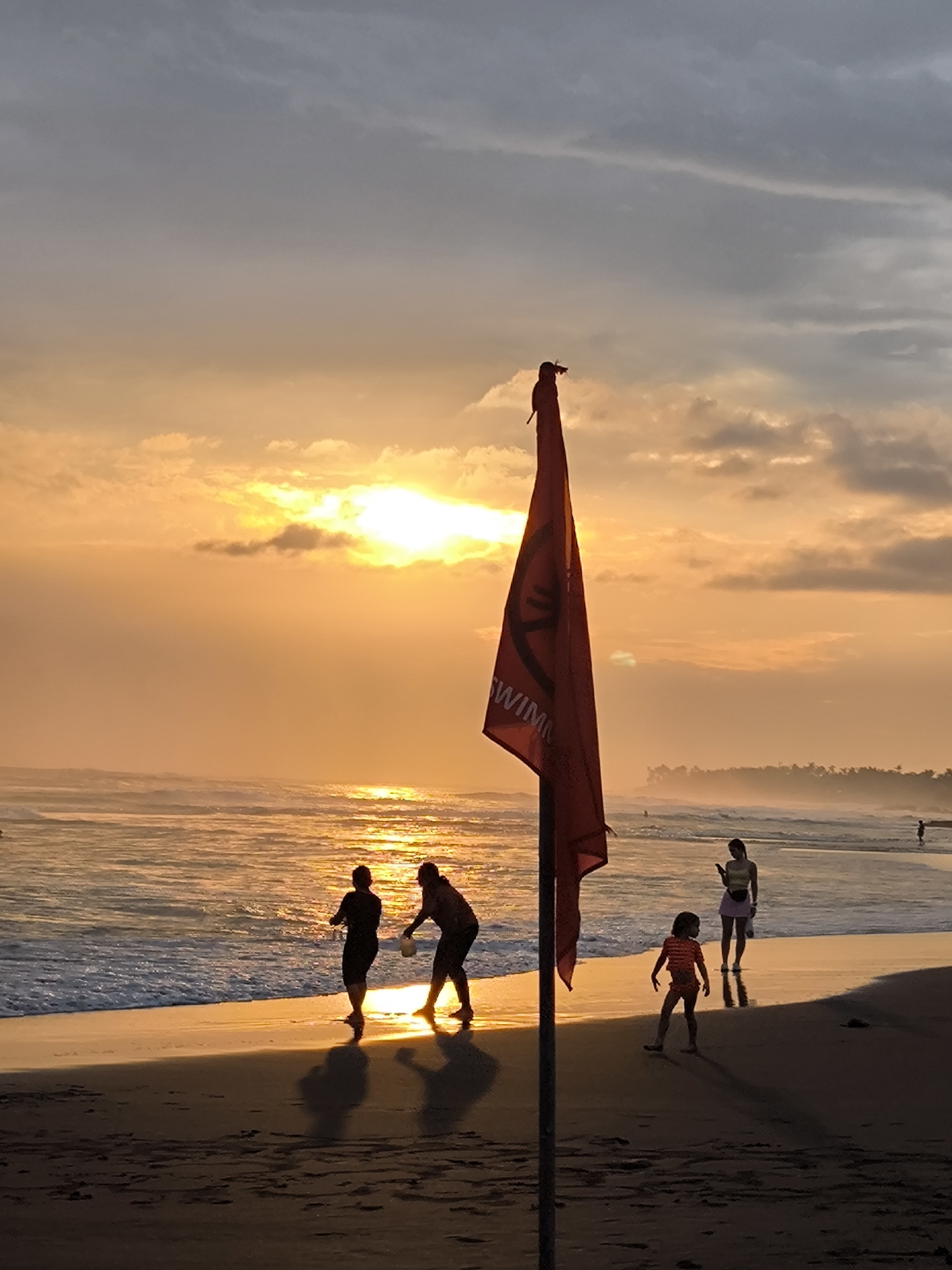 A red flag with the word "SWIMMING" printed in white is planted on a sandy beach in North Kuta, Indonesia, during sunset. The sun, low on the horizon, casts a warm orange glow across the sky and reflects off the ocean. Several people are visible on the beach, some wading in the shallow water and others walking along the shore. The silhouettes of these individuals are cast against the bright sky. The waves are gentle, lapping at the edge of the sand. The atmosphere is serene and peaceful.