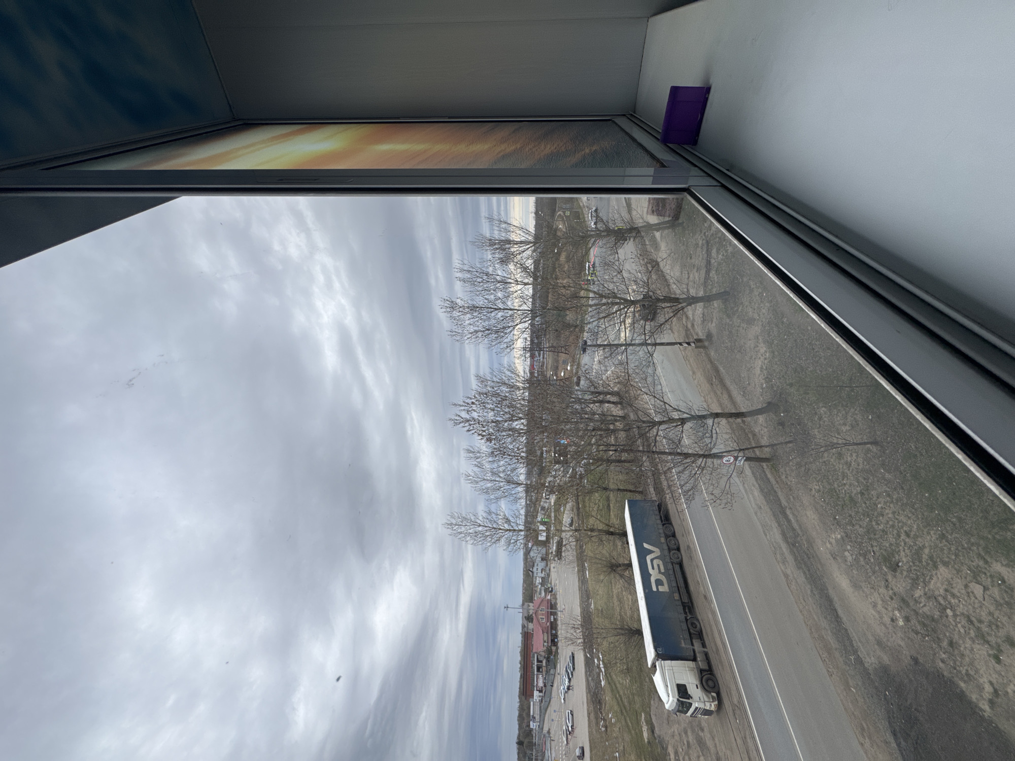 View from a window, possibly an upper story of a building, showing a street scene in Kladovitsy, Russia. Overcast sky with grey clouds. Bare trees lining the road. A large DSV-branded truck is driving on the road. Parking lot with several cars. Purple object visible on the inside window ledge.