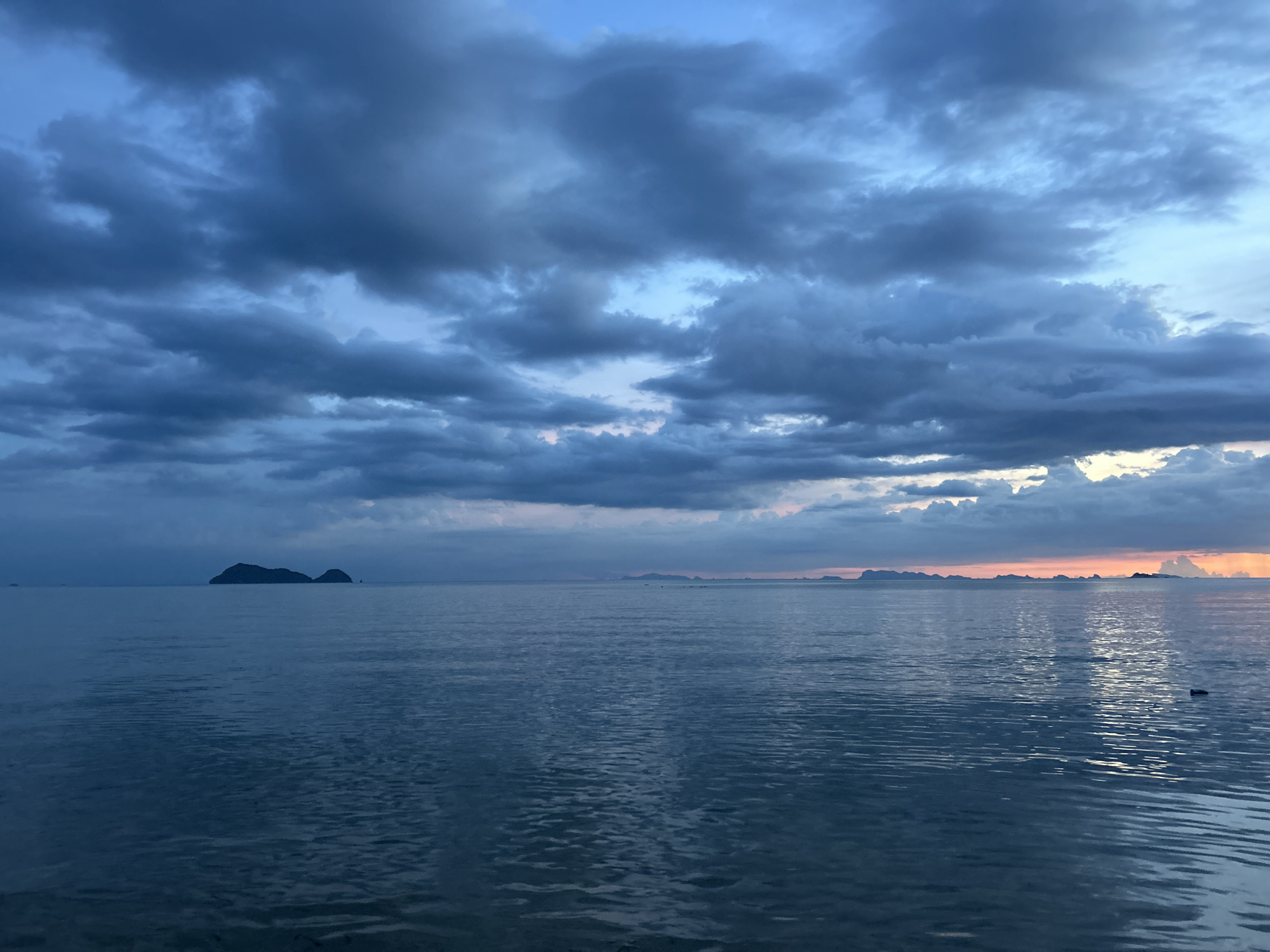 A wide shot captures a serene seascape at dusk, with dark, brooding clouds dominating the sky. The ocean reflects the muted blues and purples of the twilight, with gentle ripples creating a textured surface. In the distance, several islands are silhouetted against a faint horizon line. The foreground shows the water in greater detail, with small waves lapping. The scene is devoid of human presence. The location is identified as Tambon Ko Phangan, Thailand.