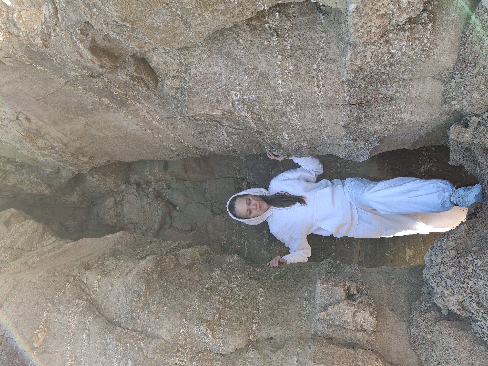 A woman wearing a white hooded sweatshirt and white pants is lying in a narrow crevice between two large rock formations. Her eyes are closed. The location is unclear, but it is in the city of Chaikovsky, Russia. The environment appears to be outdoors in a rocky, natural area. There are no other identifiable people or objects in the scene. The action is the woman lying in the crevice.