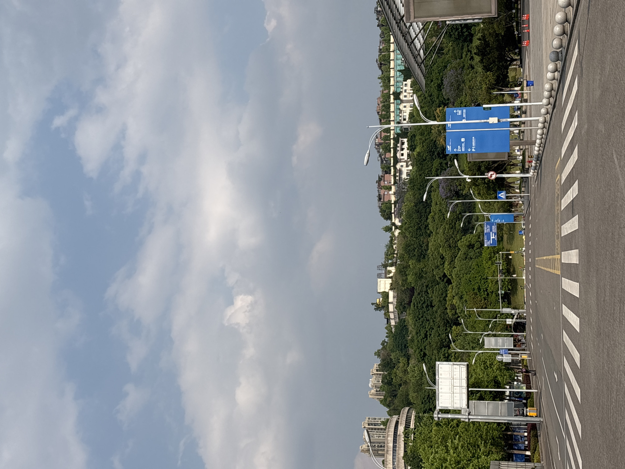 The image depicts a view in Liangjiang, China, likely along a roadway with prominent directional signage. The sky is partly cloudy. There is a mix of residential and commercial buildings in the background, interspersed with dense green trees. The roadway itself has white and yellow striped crosswalk markings. Along the side of the road are a series of streetlights and blue directional signs. A line of spherical bollards separates the walkway from the roadway.