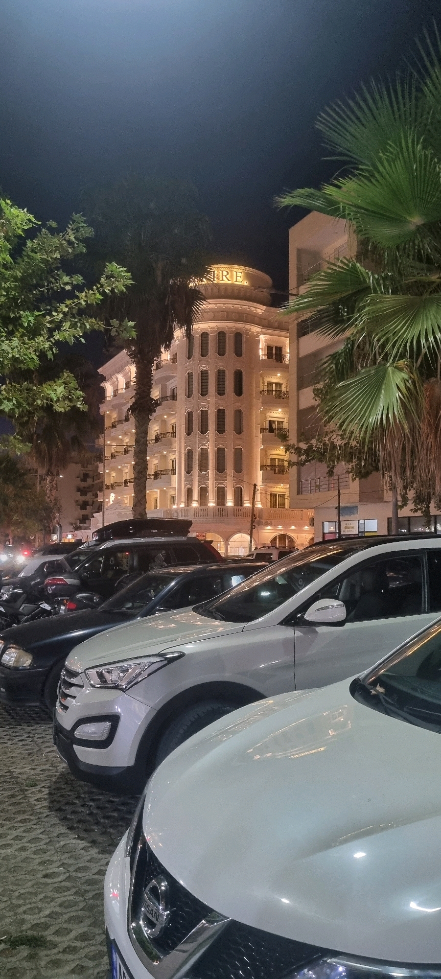 The image shows a nighttime scene in Bashkia Durrës, Albania. In the background, a multi-story building with ornate architecture and illuminated windows is visible, featuring the word "EMPIRE" in illuminated letters on its rounded top. Palm trees and other foliage frame the building. In the foreground, several cars are parked. These include a white SUV, a black SUV with a roof box, and a black car. The ground appears to be paved with interlocking stones. The lighting suggests it is a well-lit urban area. There are no people visible in the frame.