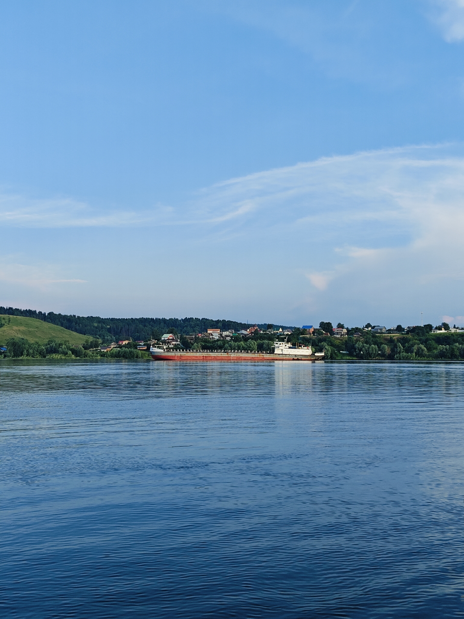 This is a landscape photograph taken from a body of water, likely a river, looking towards a shoreline. A long, flat-bottomed barge or cargo ship, primarily red and white, is docked or moored along the shore. The barge has a white superstructure and appears to be laden. The shoreline is lined with a variety of buildings, indicative of a settlement, possibly the city of Noviy, Russia. These structures are nestled amongst lush green trees and rolling hills. The water in the foreground exhibits gentle ripples, reflecting the clear blue sky with scattered wispy clouds. The overall impression is a peaceful, daytime scene of a riverside community.
