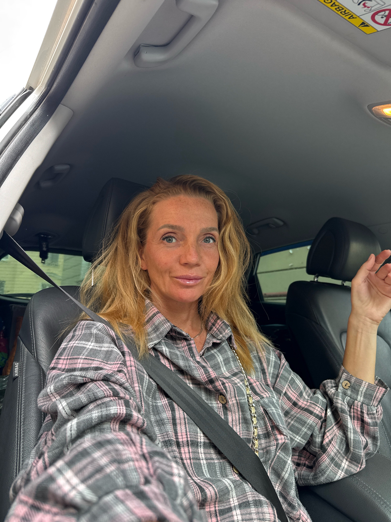 A blonde woman with freckles sits in the driver's seat of a car, looking at the camera. She is wearing a grey and pink plaid shirt and a seatbelt. Her left hand is visible, resting on her lap, and her right hand is raised slightly, with fingers extended. The interior of the car is visible, including black leather seats, the dashboard, and the rearview mirror. An "Airbag" warning label is visible on the sun visor. The background outside the car is blurred but appears to be an urban environment.
