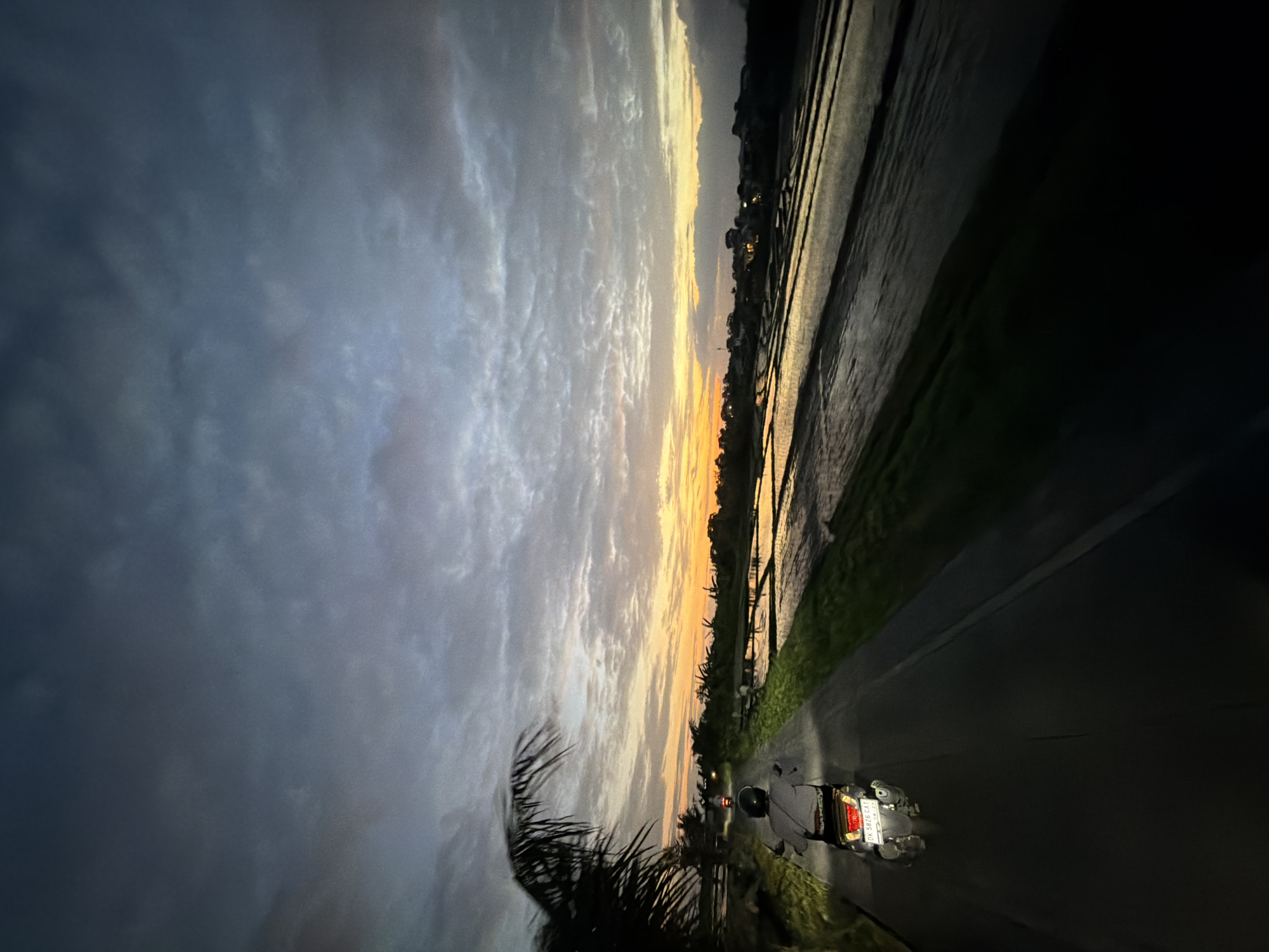 A motorcycle is parked on the side of a road in North Kuta, Indonesia, next to rice paddies. The sky is filled with clouds and a sunset is visible on the horizon. A palm tree is in the foreground.