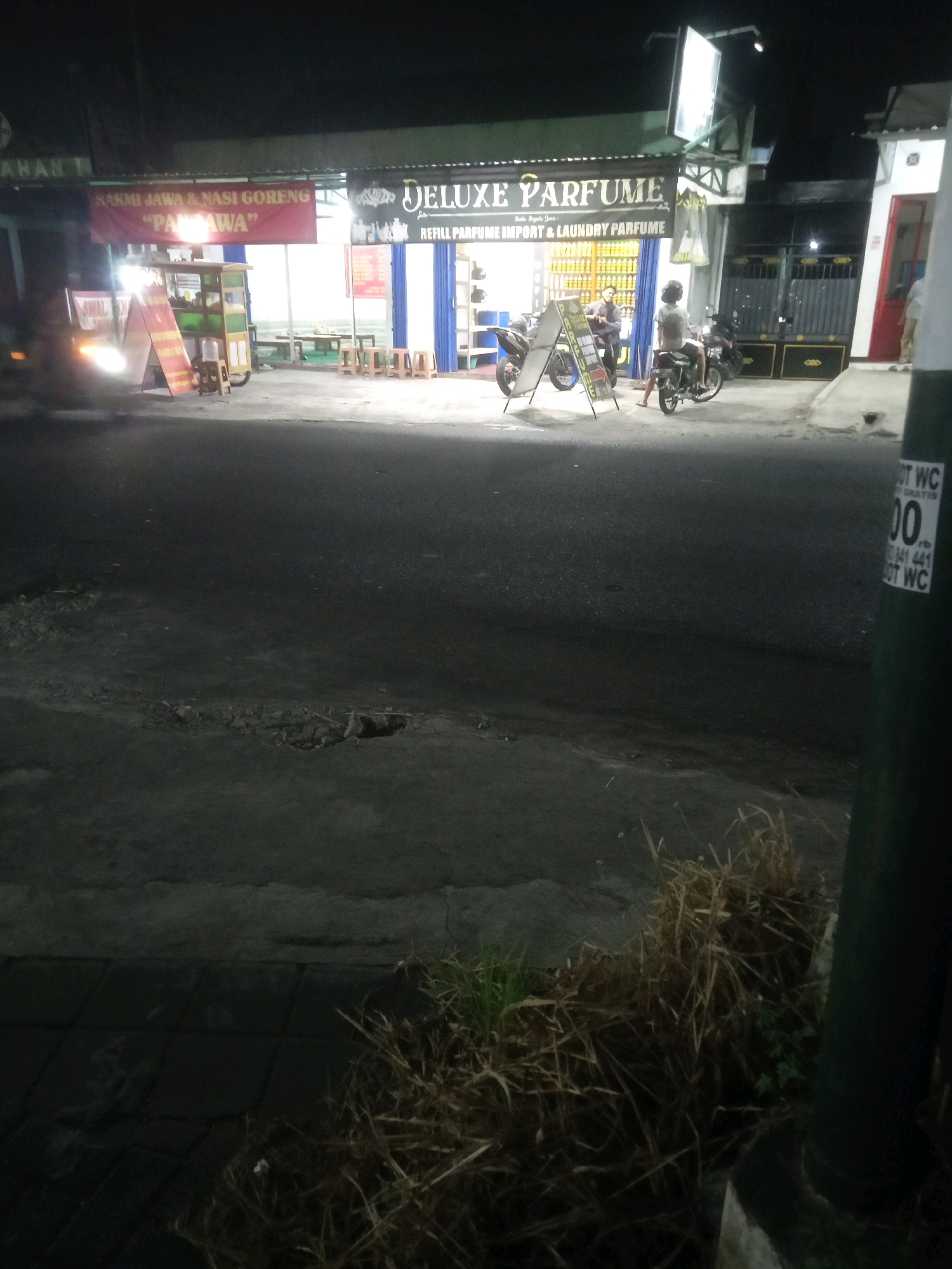 The image captures a night scene in front of a business establishment on a street in Trihanggo, Indonesia. A red banner with white text reads "SAKMI JAWA & NASI GORENG 'PA PA WA'". To the right of the banner, a sign for "DELUXE PARFUME REFILL PARFUME IMPORT & LAUNDRY PARFUME" is visible, with bottles of perfume displayed behind a blue shuttered window. Two motorcycles are parked outside, and two individuals are near them. One person is sitting on a motorcycle, wearing a helmet, and another person stands nearby. The street in the foreground is paved and appears to be a typical urban road with some debris. A dark pole with stickers, one of which indicates "WC", is present on the right side of the image. Dried grass and vegetation are visible along the edge of the street. The lighting suggests it is nighttime, with artificial lights illuminating the storefront and street.