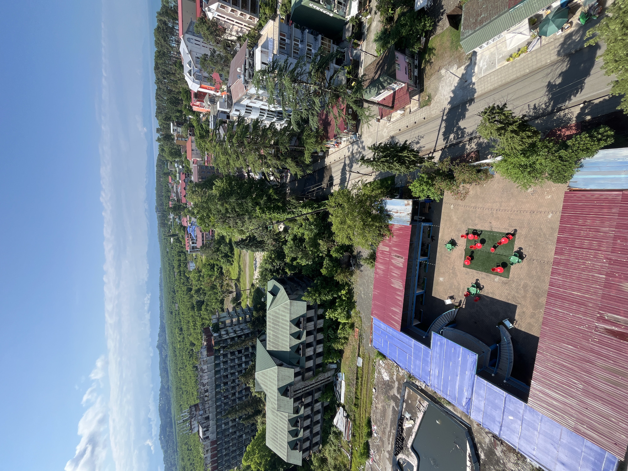 This is an aerial view of a coastal town in Ureki, Georgia. The scene is bathed in bright daylight under a clear blue sky with scattered wispy clouds. The image captures a mix of urban development and lush greenery, with the sea visible in the distance on the left.
In the foreground, we see the rooftops of several buildings, some with red corrugated metal roofs and others with blue paneling. There's a paved area with a small playground featuring red and green seating. Further into the scene, a row of buildings runs along a road lined with tall, mature trees. These buildings appear to be a mix of hotels or apartment complexes, with varying architectural styles and colors, including white, light blue, and terracotta. Some of these structures, particularly towards the left and center of the image, appear to be under construction or abandoned, with unfinished facades and exposed concrete.
Beyond the built-up area, the landscape transitions to dense green foliage, characteristic of a subtropical region, with rolling hills visible in the far distance. The overall impression is a town situated between the sea and a verdant natural environment, with signs of both active development and areas that have been left incomplete. No people are clearly discernible in the image. The time of day is clearly daytime, likely late morning or early afternoon, given the strong shadows cast by the buildings and trees.
