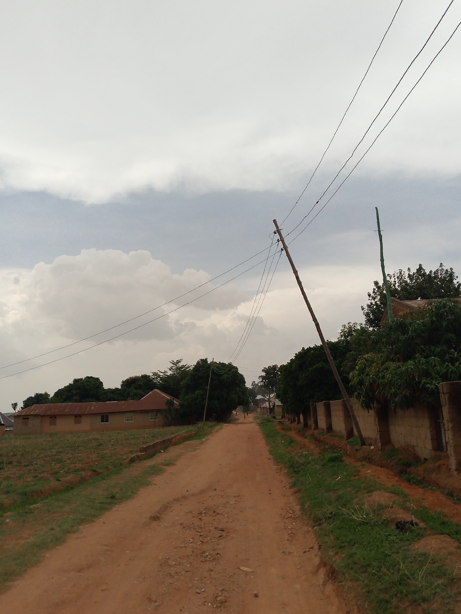 The image depicts a dirt road in what appears to be a rural or semi-rural area of Jos, Nigeria. The road is unpaved and reddish-brown in color. On the left side of the road is a field or agricultural area, with a simple, single-story building with a brown roof in the background. On the right side of the road is a concrete block wall, possibly a property boundary, with trees and vegetation behind it. An angled wooden utility pole with power lines stretches across the sky. The sky is overcast with heavy clouds. In the distance, small figures of people are visible on the road. The environment seems natural with grasses and plants lining the road edges.