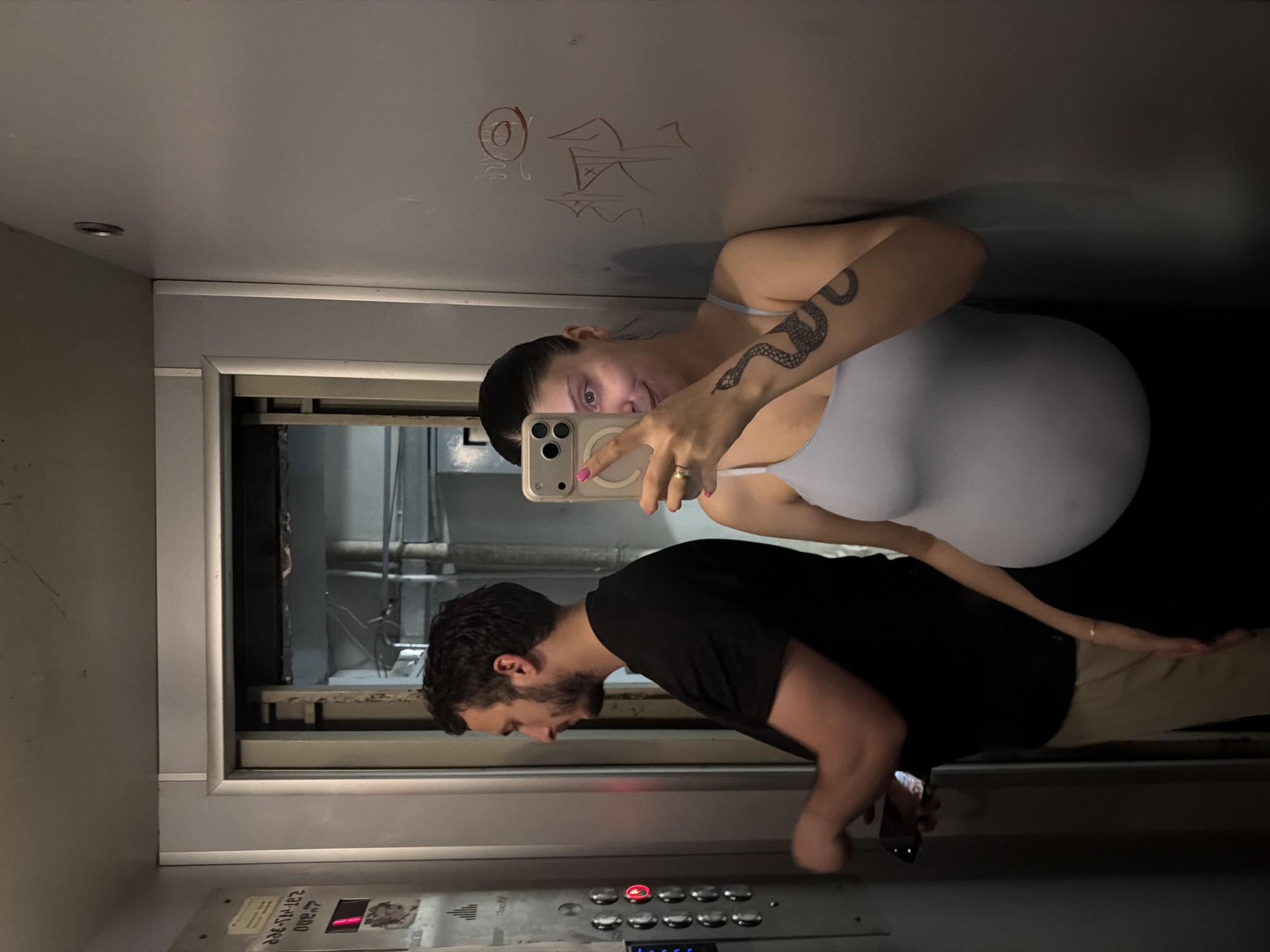 In an elevator in Tbilisi, Georgia, a man with dark hair and a beard, wearing a black t-shirt and light brown pants, stands facing the elevator control panel, holding a phone in his left hand. To his right, a pregnant woman with dark hair, wearing a light blue tank top and black leggings, leans against the elevator wall, taking a mirror selfie with her phone. She has a snake tattoo on her left arm. The elevator walls are a metallic gray, with some graffiti on the right wall. The elevator control panel on the left has illuminated buttons and a digital display showing the number "1" in red.