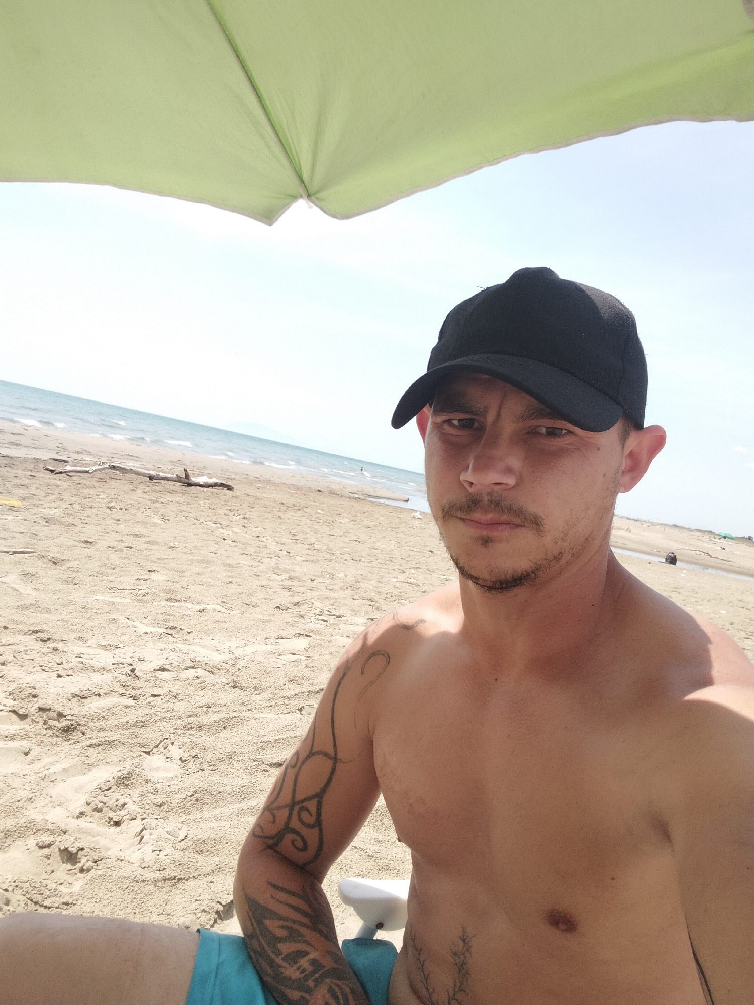 The image depicts a shirtless man with a black baseball cap on a sandy beach. He has facial hair and tattoos on his arm and torso. The background shows a beach with the sea and sky. The man is sitting under a light green umbrella. The location is likely Mondragone, Italy, given the prompt's request. The scene captures a casual beach setting with a person relaxing under an umbrella.