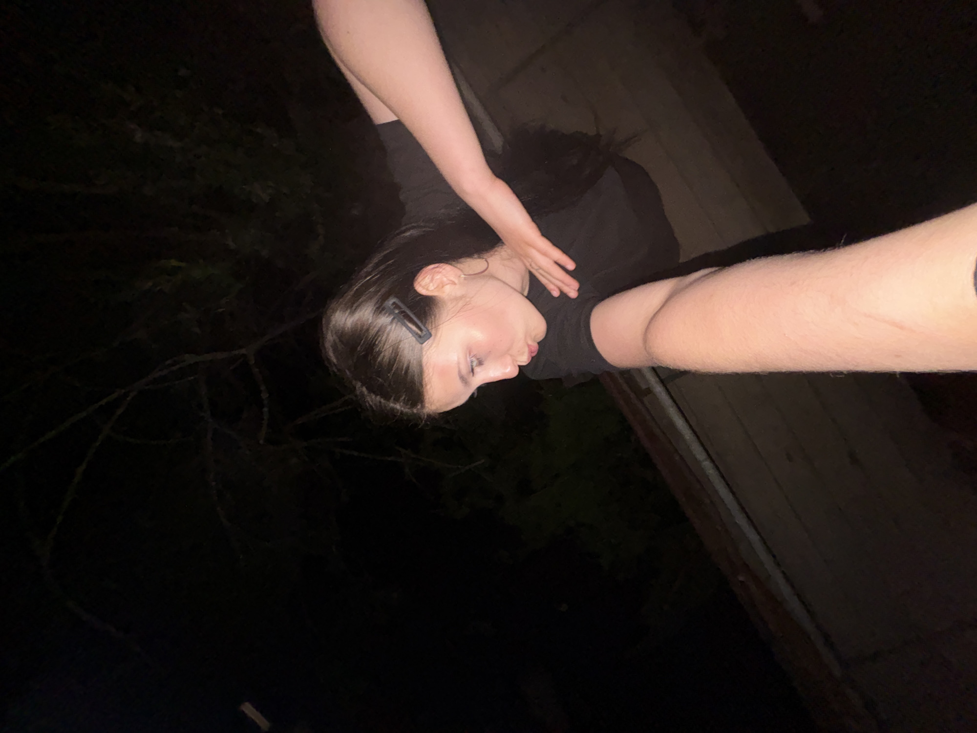 A woman is depicted in an outdoor setting, likely at night due to the darkness. She has dark hair, fair skin, and is wearing a dark top. One arm is extended upwards, and the other is outstretched towards the camera. The backdrop consists of foliage on the left and a wooden structure on the right. The image appears to be taken at an angle. There is no indication of the image being in the city of Фоки, Russia.