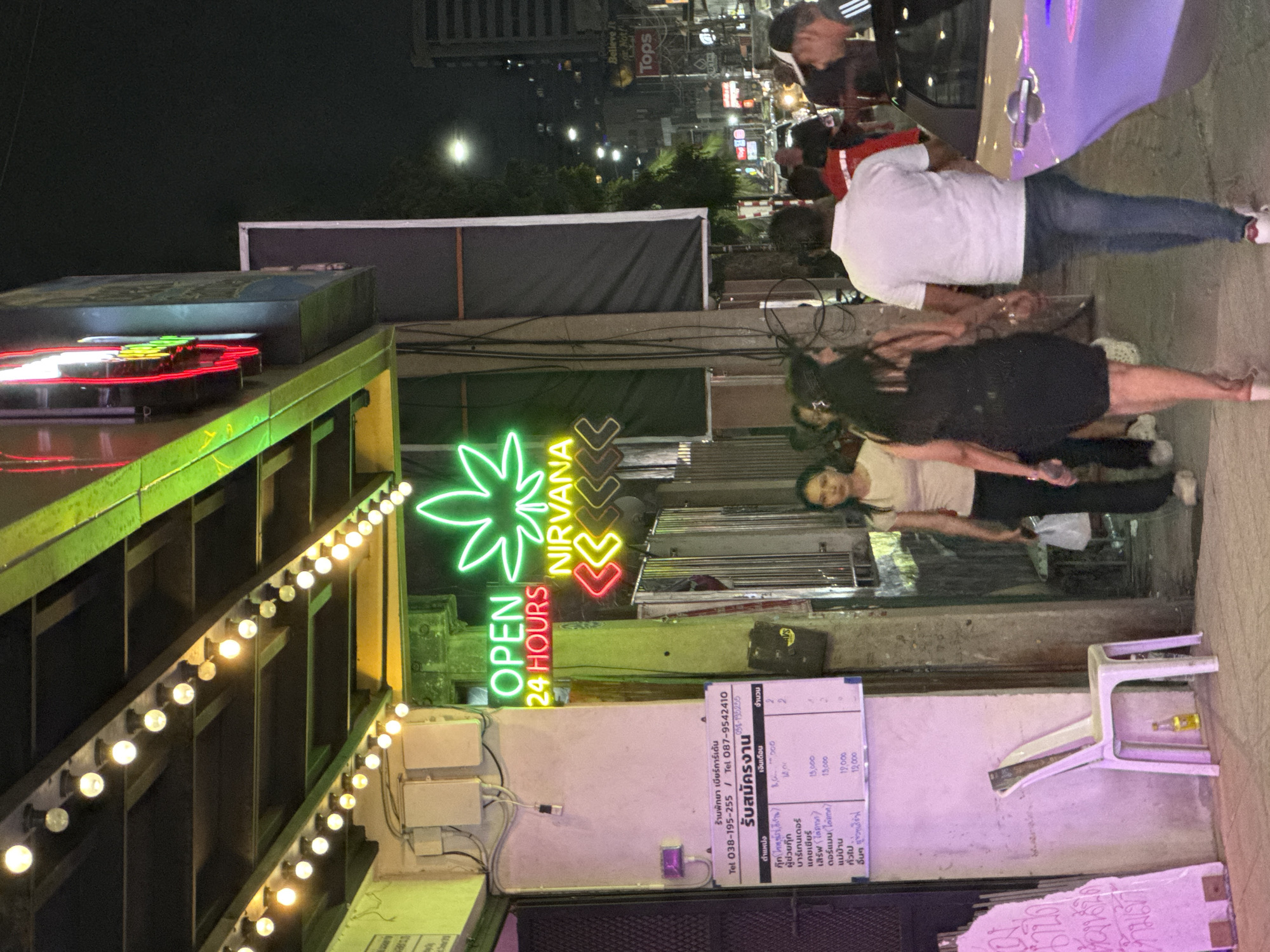 The image depicts a vibrant, illuminated street scene at night in Pattaya City, Thailand. The setting is an urban commercial area, characterized by artificial lighting and various signs.
The main subject is a prominent bright green neon sign shaped like a cannabis leaf, with the text "OPEN 24 HOURS" in green and "NIRVANA" in yellow and red neon, suggesting a local dispensary or establishment. Below this, a long string of warm incandescent bulbs outlines a structure, contributing to the evening ambiance.
Several people are visible, engaged in casual street activity. A woman in a black dress stands in the middle-ground, and another person in a white t-shirt and jeans is seen near a partial view of a light purple car. Faces are largely indistinct, and emotions cannot be discerned.
Notable objects include a white plastic chair on the pavement with a small bottle underneath. A white board attached to a building displays text in Thai, detailing job openings for "Pattaya Beer Garden" (ร้านพัทยา เบียร์การ์เด้น) along with contact telephone numbers (038-195-255 / 087-9542410). The listed positions include Assistant Cook, Bartender, Cashier, Server, Doorman, Maid, and General staff, each with an associated salary and quantity. In the background, taller buildings and additional commercial signs, such as "Believe It or Not" and "Tops," further indicate the commercial and entertainment nature of the area. The scene appears dry, with no visible adverse weather.