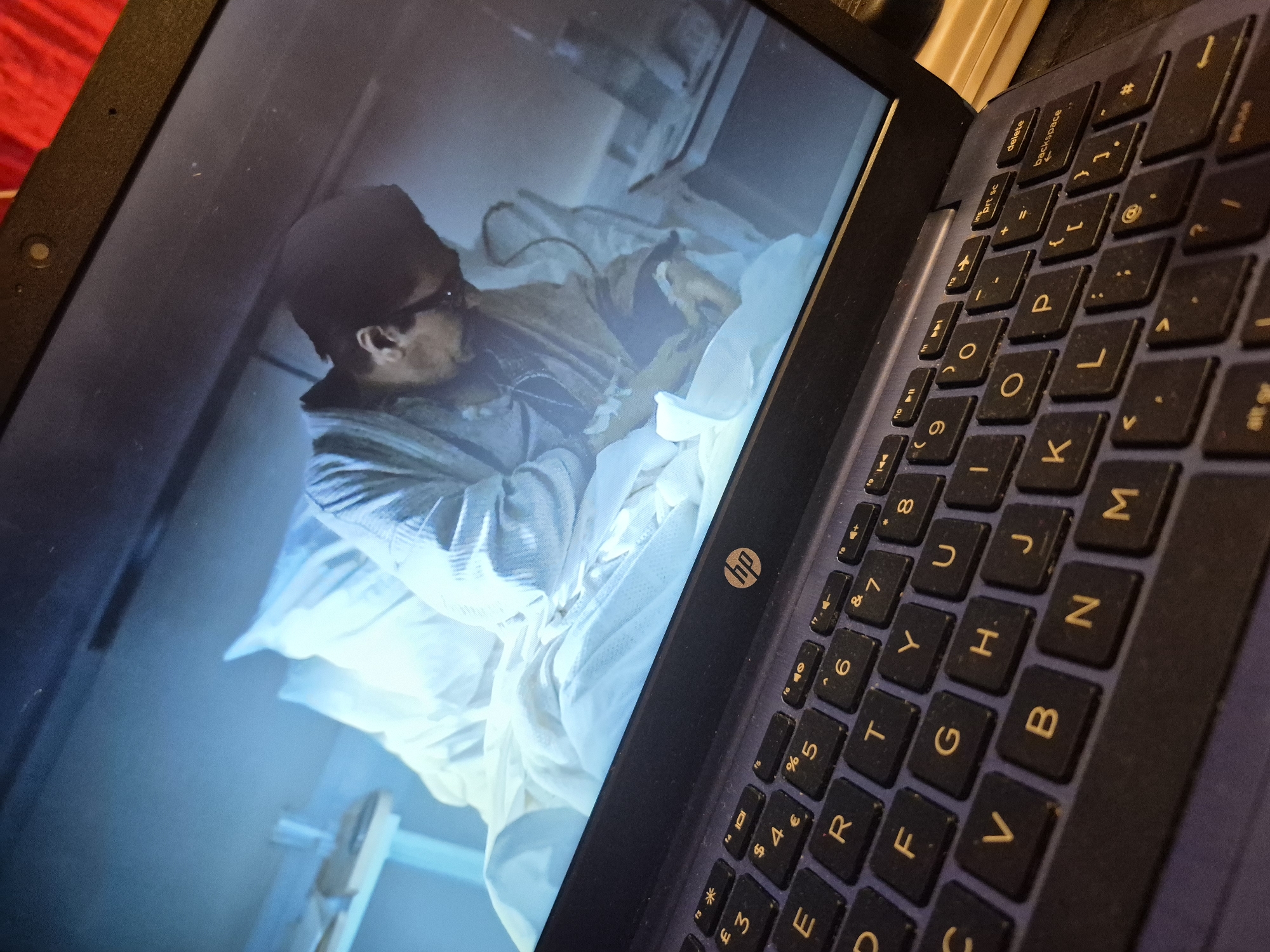 The image shows a close-up of a dark blue HP laptop. The laptop screen displays a scene from a movie or TV show, featuring a person in a hospital bed, wearing a light-colored robe, and appearing to be attended to by another person who is partially visible. The overall lighting in the on-screen scene is dim and cool-toned, with a predominantly blue hue. The keyboard of the laptop is visible in the foreground, with yellow lettering on the dark keys. The scene depicted on the screen suggests a medical or dramatic context. There is no information in the image that can be used to determine the location in the city of Sandwell, United Kingdom.