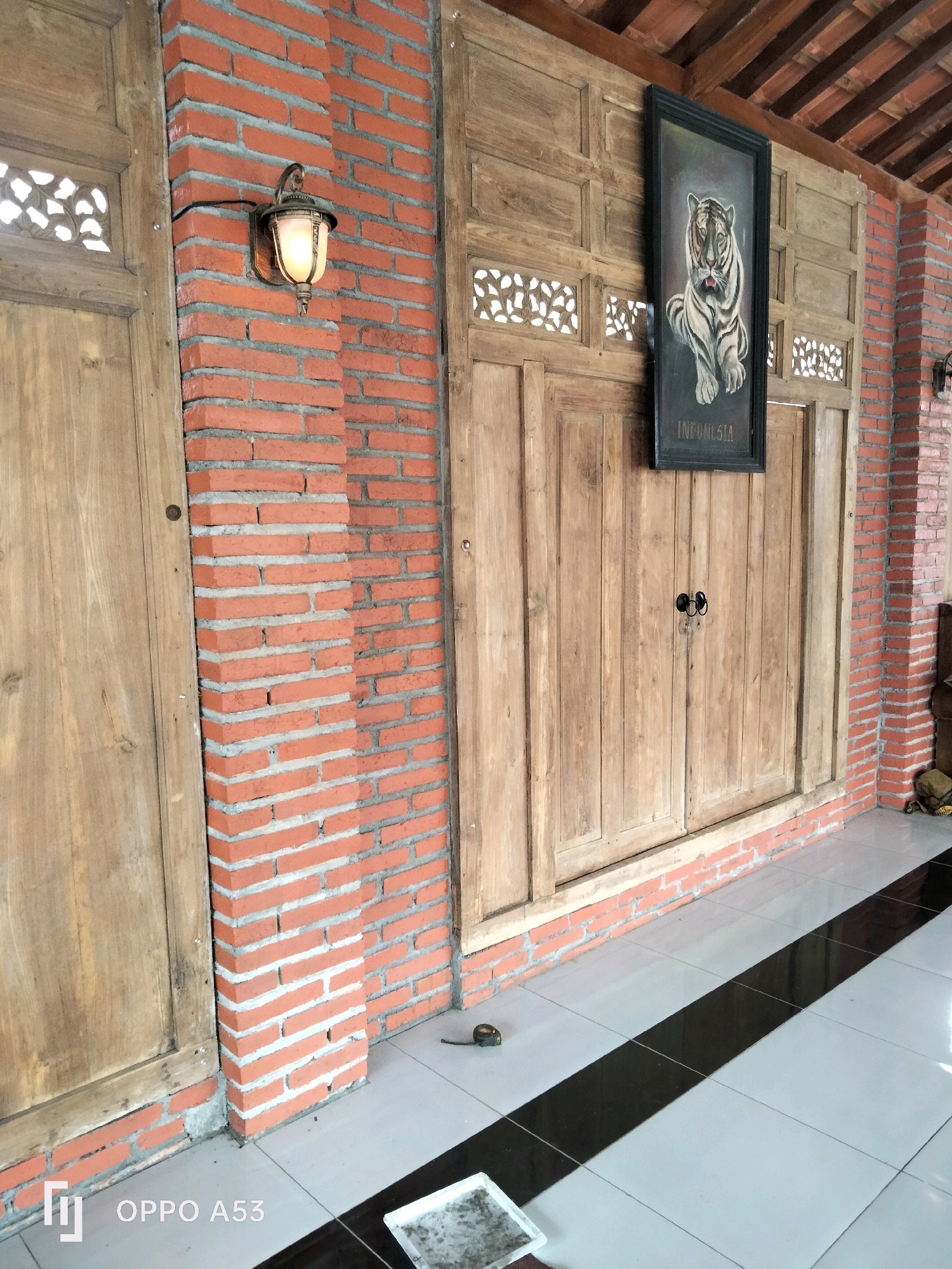 This image depicts a traditional Indonesian building entrance in Trenggalek, Indonesia. The architecture features prominent brick walls with visible mortar lines, contrasting with large wooden doors and panels. A vintage-style lantern is mounted on the brickwork, illuminating the area. Above the wooden doors, intricate carved wooden panels add decorative detail. A framed painting of a tiger with "INDONESIA" inscribed below it hangs centrally on the wooden wall. The floor is tiled with large, light-colored squares separated by a diagonal black stripe. A small measuring tape and a white tray are visible on the floor. The overall impression is one of rustic, cultural heritage.