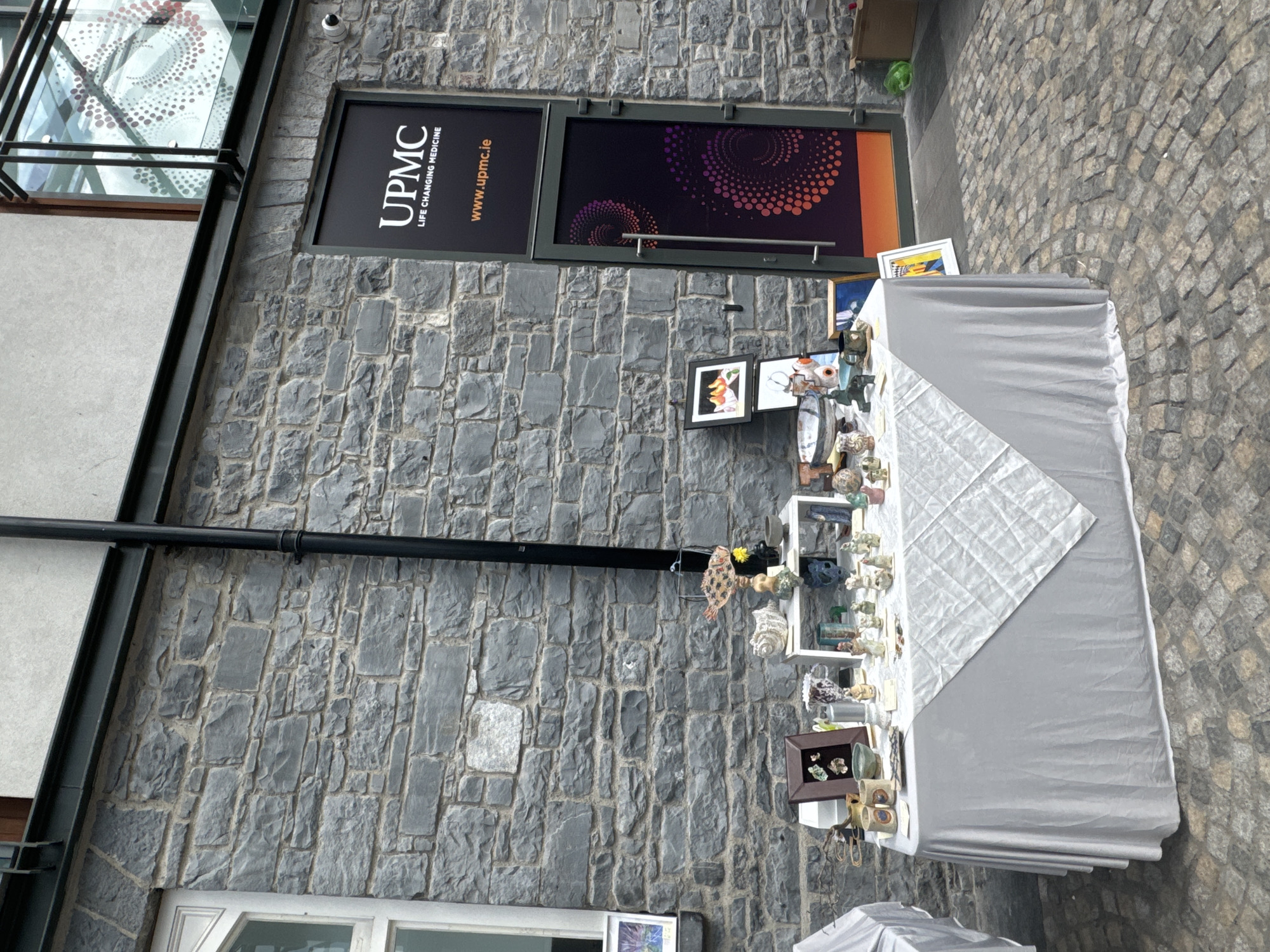 The image depicts an outdoor market stall set up against a stone-built building in Kilkenny, Ireland. A grey tablecloth drapes over a table laden with various ceramic figurines and small artworks, including what appear to be small animal figures, vases, and framed pieces. Among the items on display are two framed artworks, one depicting three yellow fruit and another with abstract elements. To the right of the stall, a glass-fronted door with a UPMC Life Changing Medicine sign is visible. The building's facade is constructed of rough-hewn grey stones, and a black downspout runs vertically along it. The ground is paved with cobblestones. There are no people visible in the immediate vicinity of the stall or the building entrance.