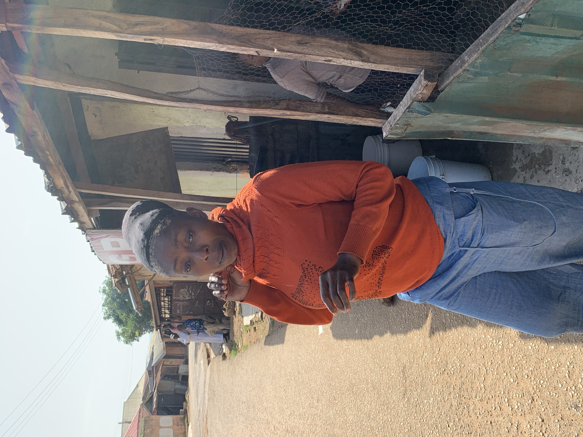 In Jos, Nigeria, a woman stands in an outdoor setting, possibly a market or residential area, with a dirt road leading into the background. She is wearing an orange turtleneck sweater with a textured design and loose-fitting blue pants. Her head is covered with a dark cloth. Her hands are near her face, with one touching her cheek. To her right, a structure made of rough wooden beams and corrugated metal roofing is visible, partially obscured by chicken wire fencing. A person in a light-colored top is partially visible behind the fencing. Two white buckets are placed on the ground near the woman. In the background, several small buildings with tin roofs line the road, and power lines are strung overhead. The lighting suggests it is daytime.