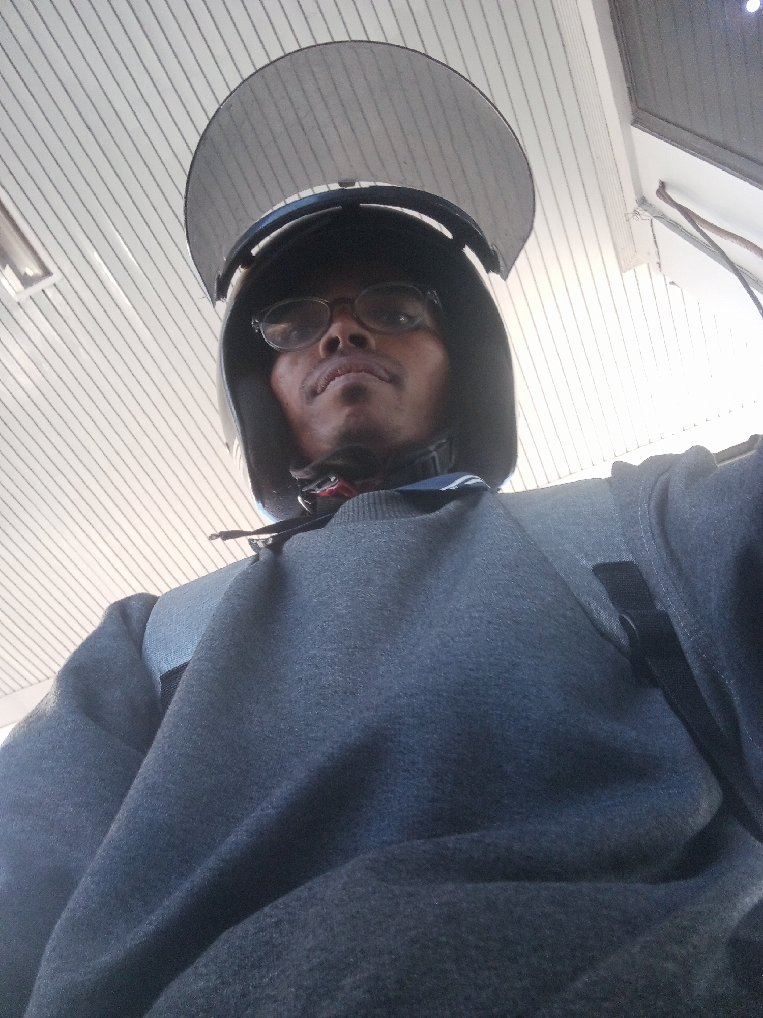 A man wearing a dark gray hooded sweatshirt and a black helmet with a clear visor is pictured from a low-angle perspective. He is wearing eyeglasses and appears to be looking upwards. The background consists of a white ceiling with parallel lines, possibly indicating an indoor setting or a covered outdoor area in Sawotratap, Indonesia. The lighting is bright, suggesting it may be daytime.