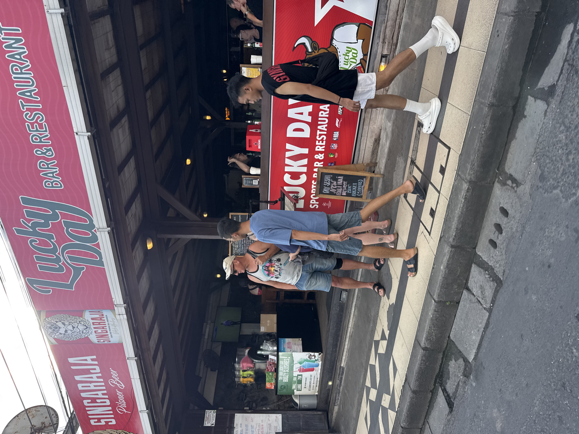 This daytime image depicts a street scene in Kuta, Indonesia, focusing on the exterior of "Lucky Day Bar & Restaurant." Three men are casually walking on the sidewalk in front of the establishment. The man nearest the camera wears a white tank top, denim shorts, a white baseball cap, and black flip-flops. Beside him, another man sports a light blue t-shirt, denim shorts, and black flip-flops. Further to the right, a third man, mid-stride, is dressed in a black t-shirt, white shorts, white socks, and white Crocs.
The bar's facade is adorned with prominent red banners. A large banner on the left reads "Lucky Day BAR & RESTAURANT" and advertises "SINGARAJA Pilsner Beer," featuring a can graphic with "KUALITAS PREMIUM ASLI INDONESIA." A red wall poster also displays "Lucky Day SPORTS BAR & RESTAURANT" alongside logos for NBA, UFC, Formula 1, and Champions League.
Promotional signs are visible, including a black A-frame chalkboard sign offering "DAY SESH!", "NEW: BALI HAI Day Lager," and "150K BUCKET (5 BEERS)." Another sign advertises "TRY OUR LUCKY DAY BOOZY SLUSHIES!" with prices: "SMALL 35K," "REG 55K," and "LARGE 75K." Inside the bar, a red BINTANG box can be seen.
The sidewalk features a distinctive black and white checkered pattern leading to grey concrete sections, adjacent to an asphalt road. The bright lighting suggests it is daytime. The overall ambiance is relaxed, indicative of a casual street in a popular tourist destination like Kuta.