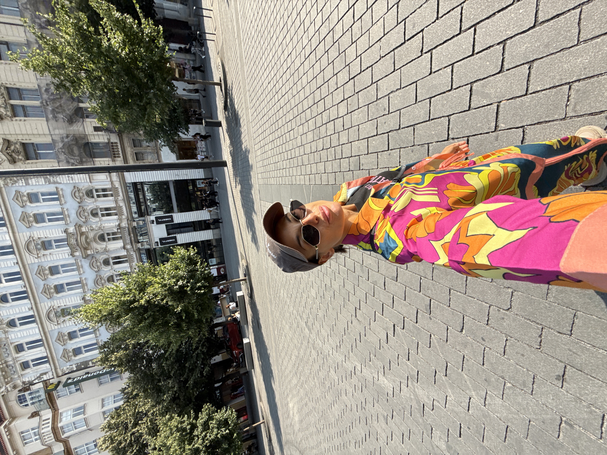 A young woman in a colorful patterned outfit and a baseball cap takes a selfie on a cobblestone plaza in Prague, Czechia. She is wearing sunglasses and her lips are slightly parted. Behind her, pedestrians stroll past historic buildings, some with intricate architectural details and balconies. Trees line the plaza, casting shadows on the ground. The scene appears to be a public square, likely in the historic center of the city, given the architectural style of the buildings. The lighting suggests it is daytime, with the sun casting strong shadows.