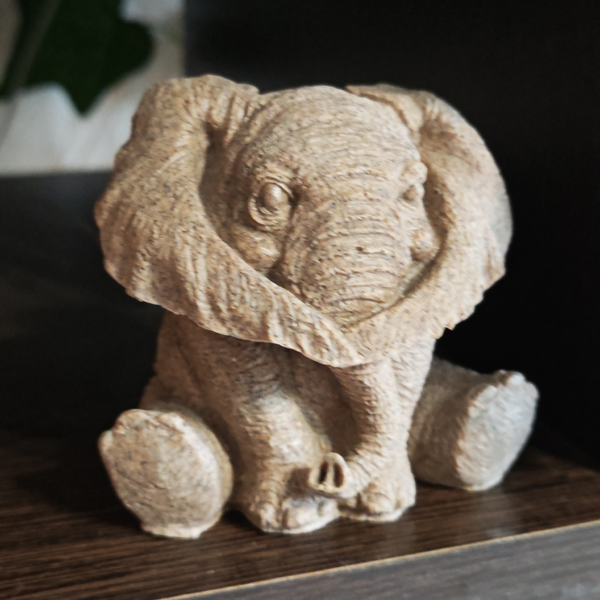 A close-up shot displays a small, tan-colored elephant figurine sitting on a dark wooden surface. The figurine is intricately detailed, showing the elephant's wrinkled skin, large ears, and small tusks. The background behind the figurine is dark and blurry, suggesting an indoor setting, possibly a shelf. No people or actions are discernible in the image. The location of the image, Kladovitsy, Russia, cannot be confirmed solely based on the visual information.