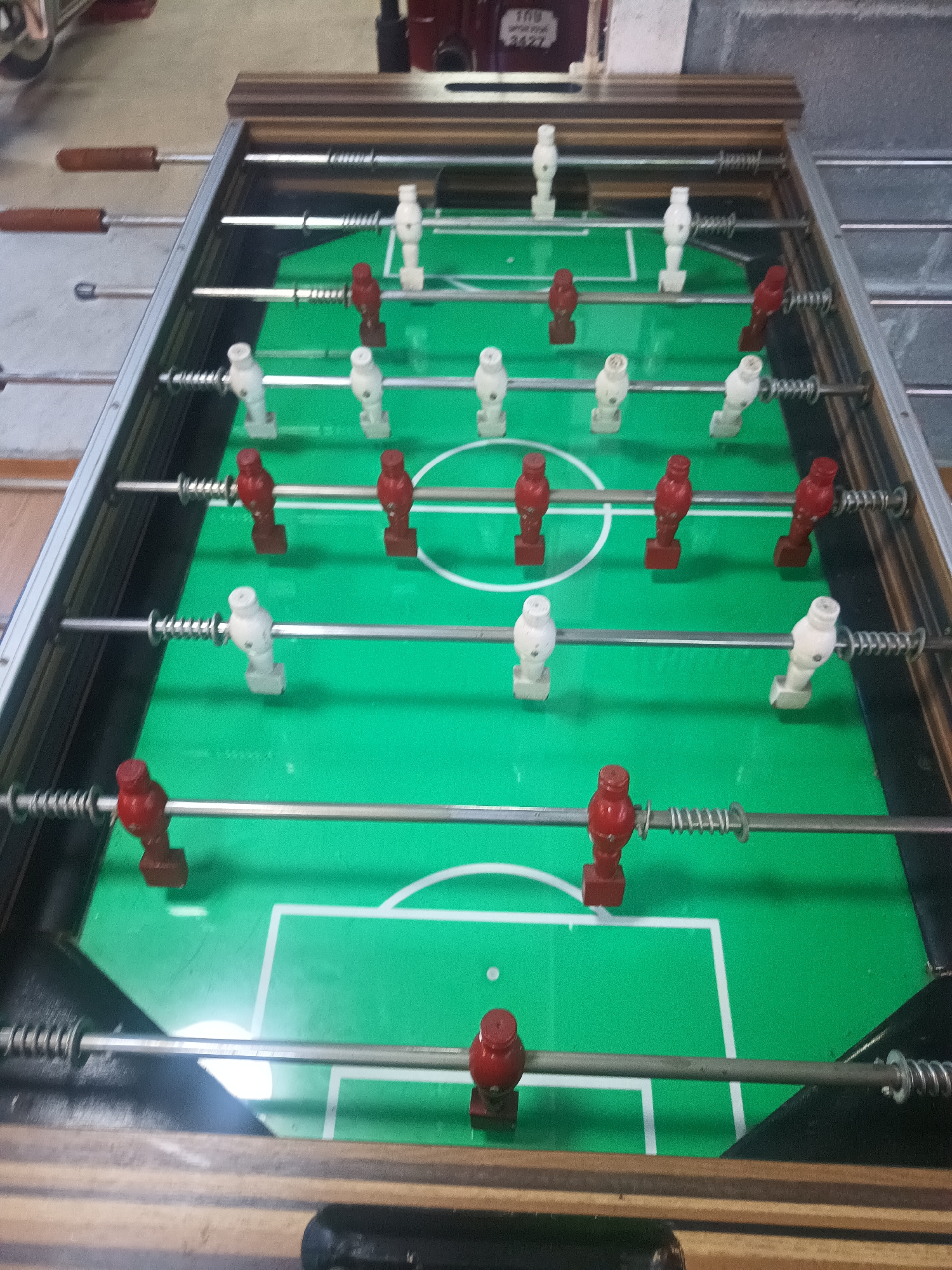 A foosball table is the central element of the image. The table is green and features red and white foosball players mounted on metal rods. The rods are attached to the sides of the table, which has a wooden frame. The setting appears to be indoors. The location cannot be determined as Surat Thani City Municipality, Thailand based on the image alone.