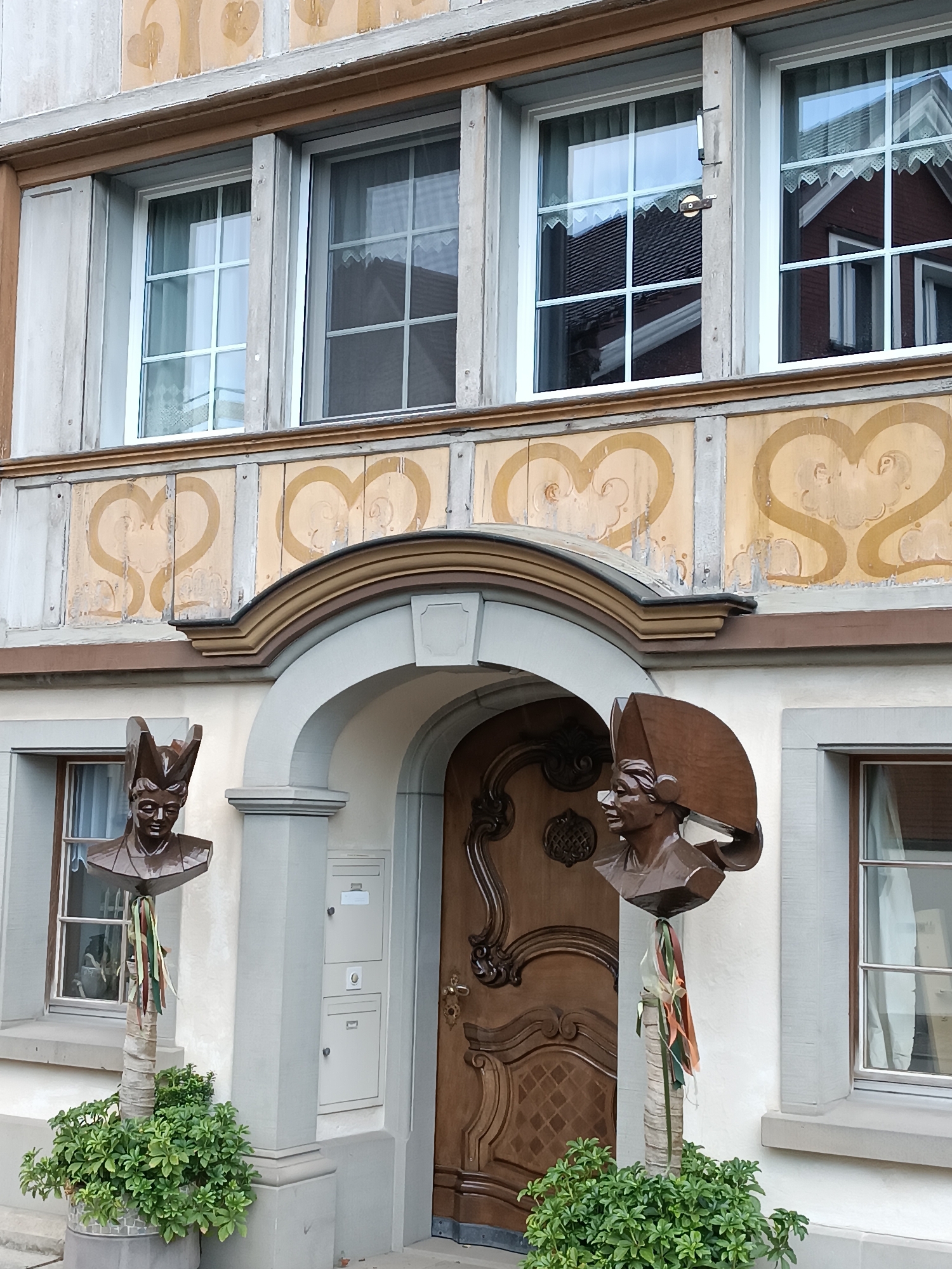 The image shows the facade of a building in Appenzell, Switzerland. The building has a light-colored stucco exterior with decorative elements in a cream and yellow palette, including heart-shaped motifs and painted wooden trim. There are multiple windows with white frames and a grid pattern. An arched entryway with a dark wooden door features intricate carving. Flanking the entrance are two busts mounted on wooden poles, each adorned with colorful ribbons. The bust on the left is a dark bronze color and depicts a figure with a pointed hat, while the bust on the right is made of carved wood and features a similar hat. Green plants in pots are situated at the base of the busts. The overall scene suggests a historic or traditional architectural style characteristic of the region.