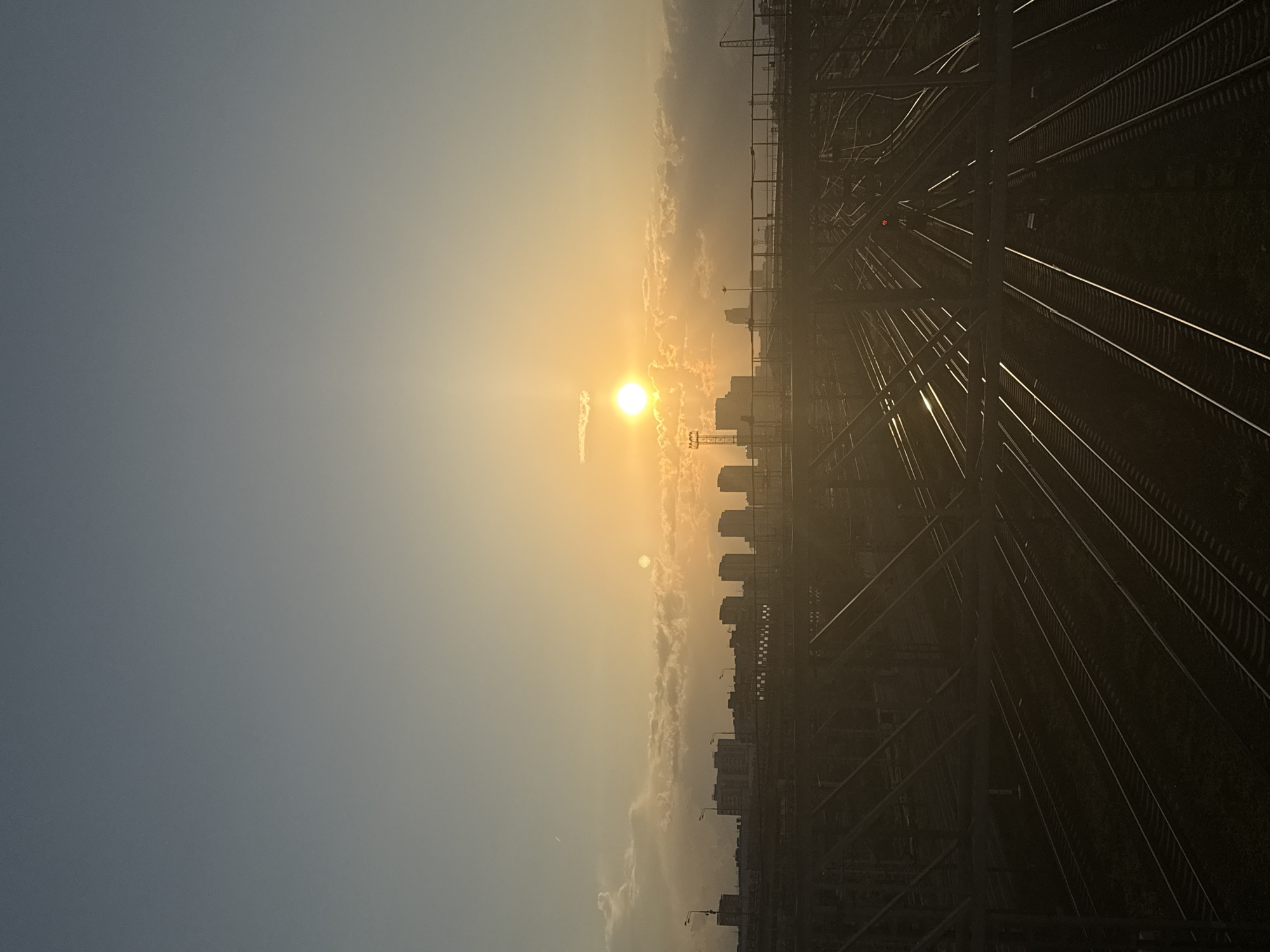 The image captures a sunset over the railway tracks near Moscow, Russia. The sun is positioned in the center of the frame, surrounded by a hazy sky and some clouds. The city skyline is visible in the background with buildings and structures silhouetted against the sunlight. Numerous railway tracks dominate the foreground, with some reflection of the setting sun. The environment is urban, with transportation infrastructure being the main focus. There is no evidence of people present in the scene.