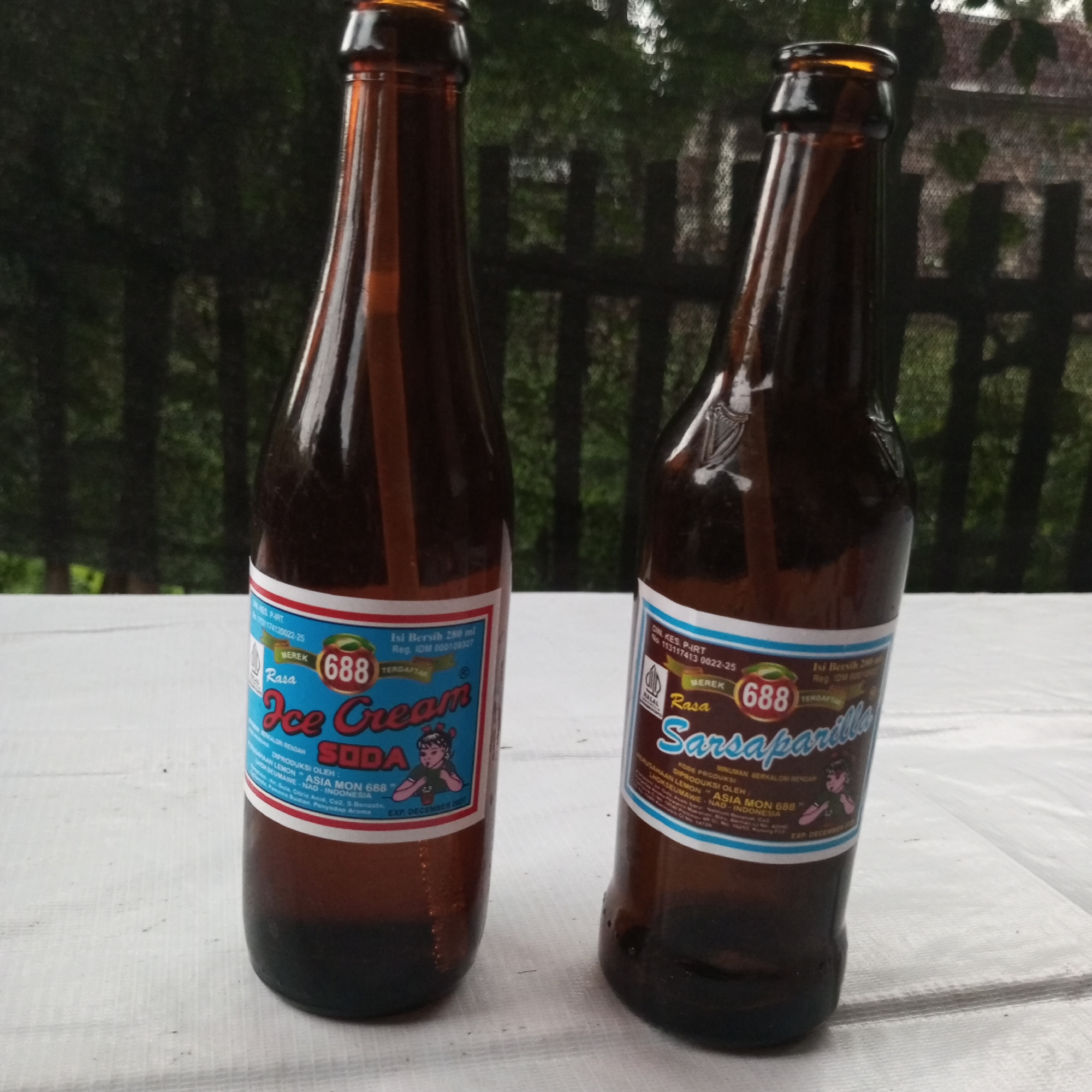 Two brown glass bottles, each containing a dark liquid and featuring a distinct blue-and-white label, are positioned upright on a white, textured surface. The scene is set outdoors in Teupinjok, Indonesia, with a soft-focus green background of foliage and indistinct dark structures.
The bottle on the left, identified by its label as "Rasa Ice Cream Soda," belongs to the "688" brand. Its label specifies a net content of 280 ml, a registration number (Reg. IDM 000109327), and indicates it was produced by "Perusahaan Lemon 'Asia Mon 688'" in Lhokseumawe, NAD, Indonesia. The label also displays a cartoon image of a girl drinking from a straw and an expiration date of "EXP. DECEMBER 2007."
The bottle on the right, also from the "688" brand, is labeled "Rasa Sarsaparilla." Its label includes a "HALAL" certification logo, a DIN. KES. P-IRT number (113117413 0022-25), and notes it as a "MINUMAN BERSODA BERKALORI RENDAH" (low-calorie carbonated drink). A cartoon image of a boy drinking from a glass is present, and like its counterpart, it states an expiration date of "EXP. DECEMBER 2007."
Both bottles exhibit consistent branding and production information. The white surface they rest upon shows slight creases. The background consists of blurry green vegetation, likely trees, providing depth to the image, with darker, out-of-focus vertical elements suggesting a distant fence or building structure. Natural light illuminates the bottles from the front-right.