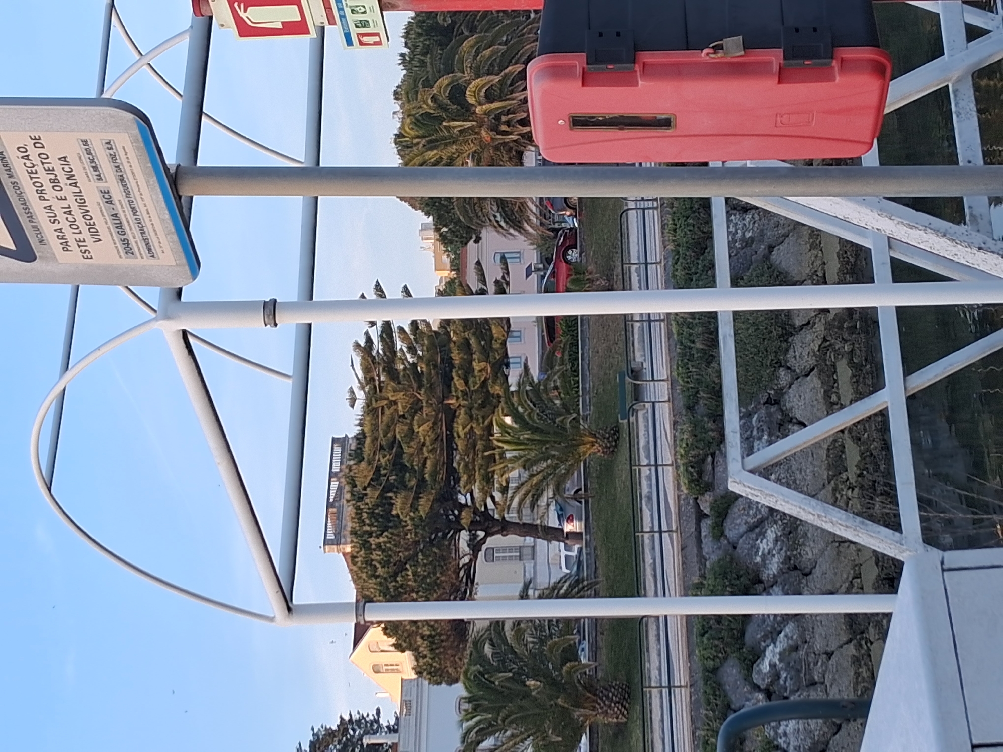 This is a daytime, low-angle outdoor shot taken in Figueira da Foz, Portugal. The scene includes a sign that reads "INCLUI PASSADIÇOS MARINA PARA SUA PROTEÇÃO, ESTE LOCAL É OBJETO DE VIDEOVIGILÂNCIA." There is also text indicating "2045 GALIA-ACE 84,58,5C,50,GE ADMINISTRAÇÃO PORTO FIGUEIRA DA FOZ, S.A."
The view includes trees, buildings, a red car, and a paved walkway alongside grassy areas. There's a red and black toolbox attached to a white metal structure. The sky is visible in the background.