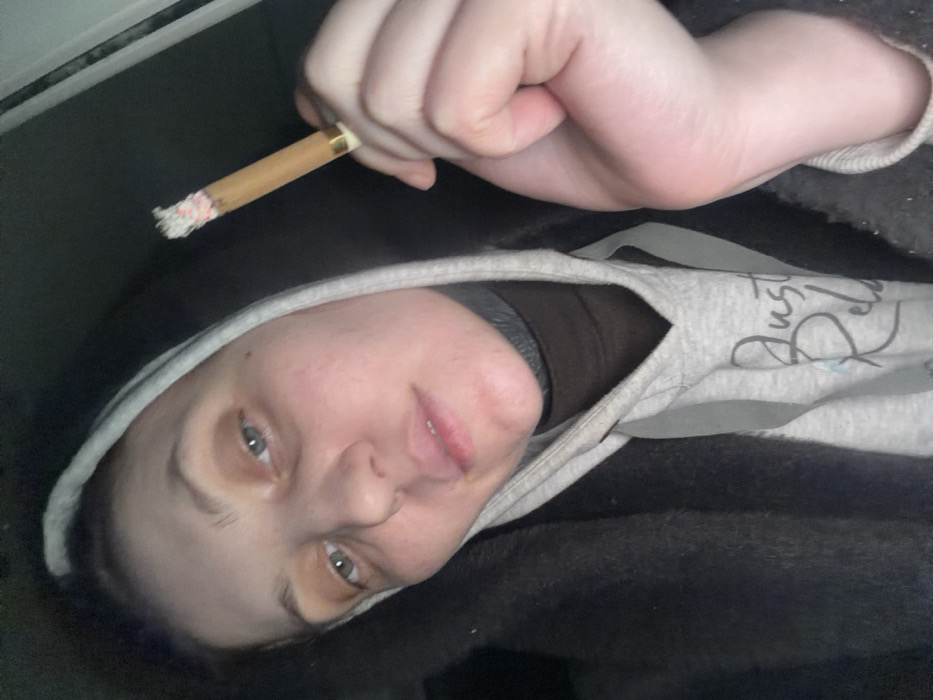 A person in a gray hoodie lies on a dark surface. The person is holding a lit cigarette in their hand. The person has light skin, eyes, and hair, and is looking at the camera. The hoodie has a phrase “Just Relax” written on it. The background is dark and blurred.