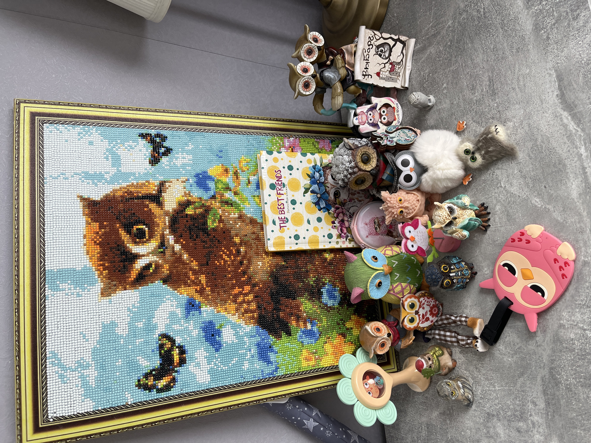 The image depicts a collection of owl-themed items arranged on a gray, textured surface. These items include: an owl diamond painting in a gold frame, a book titled "THE BEST FRIENDS" (in a non-English script) with a yellow polka-dot cover, multiple owl figurines made of various materials (e.g., plastic, ceramic, fabric), an owl-shaped baby toy, an owl-themed scroll, and other owl-related trinkets. The objects are clustered together with some items overlapping others. The location is not evident from the image; there is no information to link the scene to Shushary, Russia.