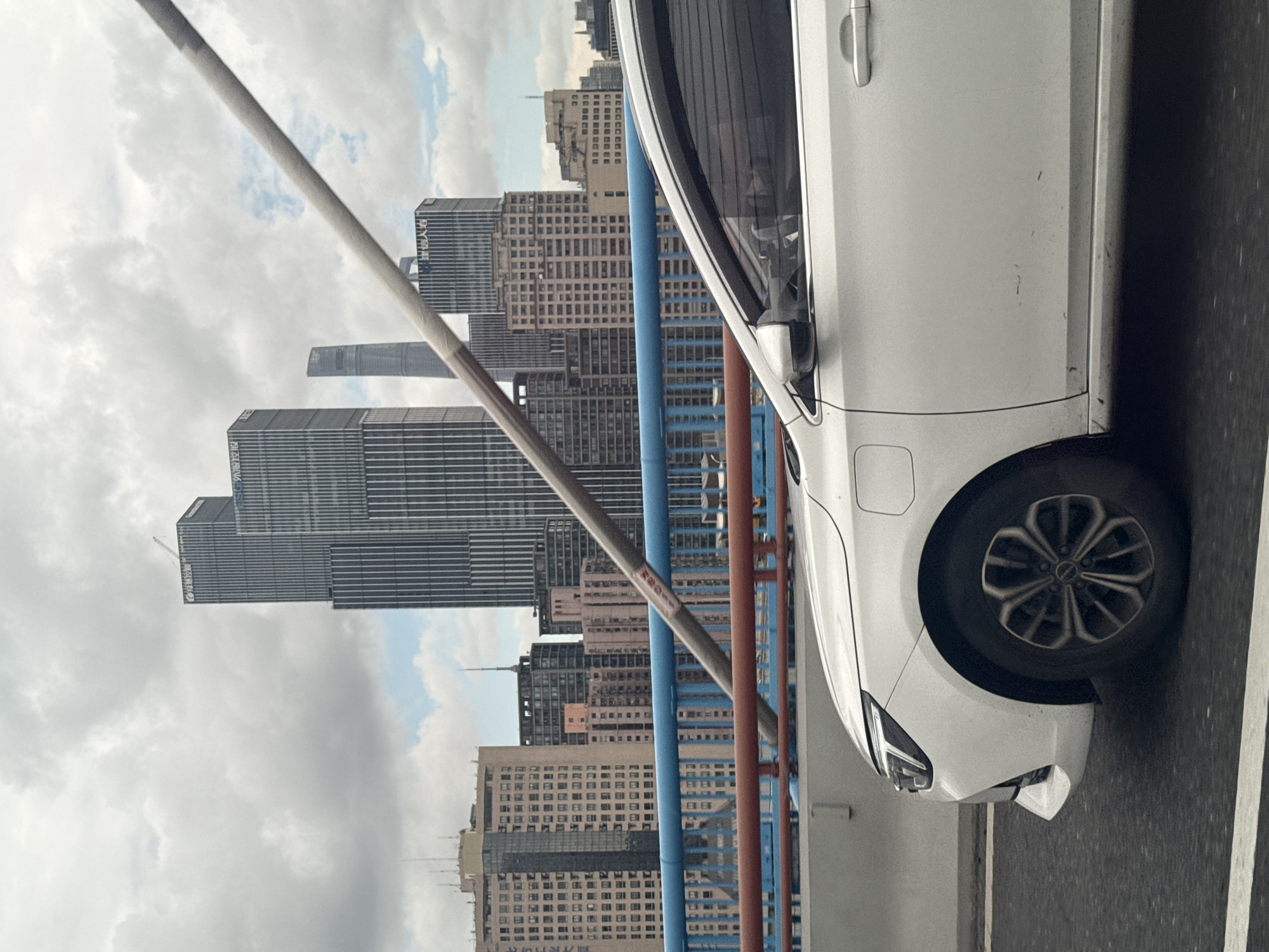 A white car drives on a bridge in Shanghai, China, with a city skyline visible in the background. The skyline features modern skyscrapers of varying heights and architectural styles, some with vertical lines and glass facades. The sky is overcast with clouds. The bridge has a blue and red railing.