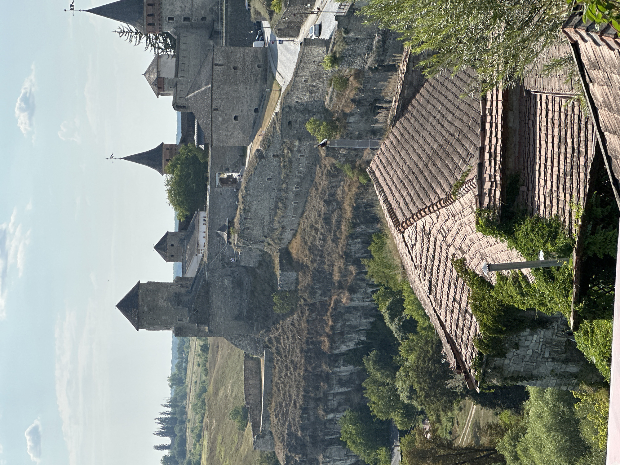 The image depicts a panoramic view of the Kamianets-Podilskyi Castle in Ukraine, a historic fortress situated on a rocky island connected to the mainland by a bridge. The castle, constructed from stone, features several towers and defensive walls. In the foreground, a tiled roof of a building is visible, partially covered in ivy. The background shows a lush green landscape with trees and rolling hills under a partly cloudy sky. A road with a few cars is visible winding its way up towards the castle.