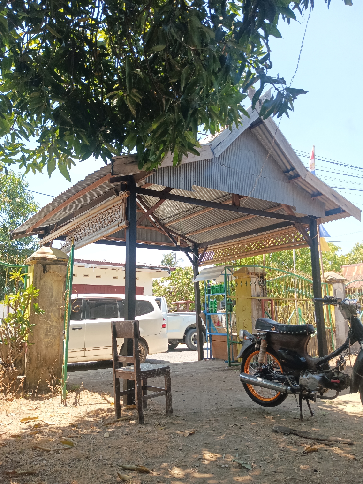 This image depicts an outdoor scene in Pitue, Indonesia. In the foreground, a weathered wooden chair sits on dry, dusty ground, partially shaded by a nearby tree. Behind the chair, a dark-colored motorcycle is parked. The background features a corrugated metal roof structure supported by dark metal poles, likely a covered outdoor area or carport. A white SUV and a white pickup truck are visible parked under the structure. Further back, a fence and some buildings can be seen, along with lush green trees and a bright blue sky. The overall impression is of a rural or suburban setting under strong sunlight.