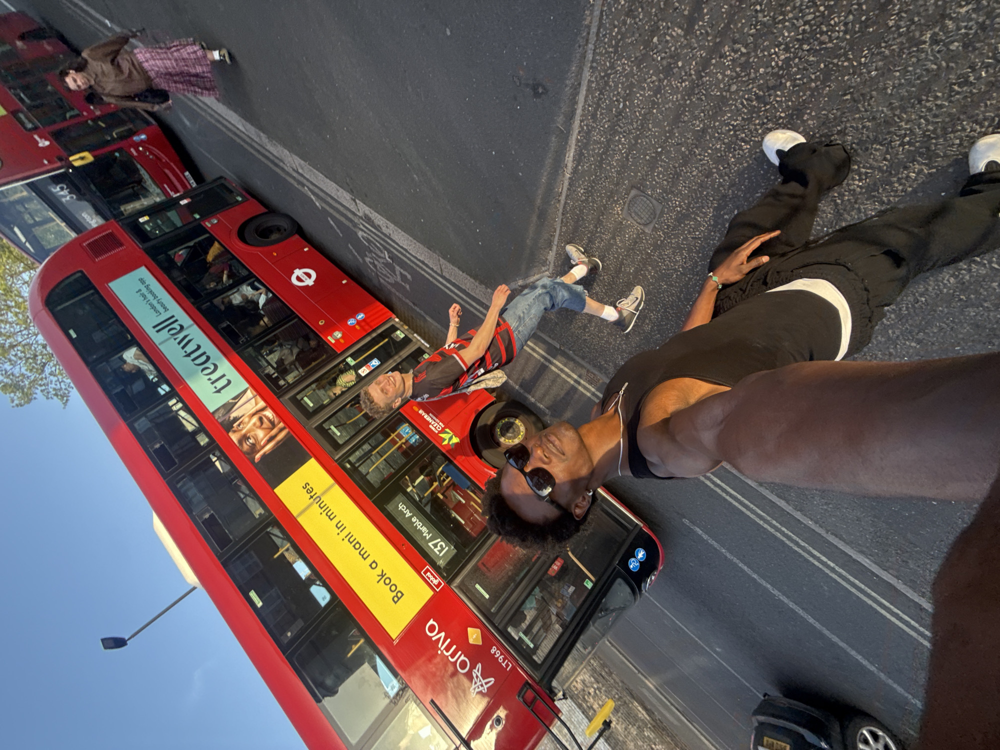 A ground-level selfie taken on a street in London, United Kingdom. A black man with sunglasses and dark hair is angled towards the camera. To his right, there is another man lying on the ground, wearing short jean shorts, a black-and-red striped shirt, and sneakers. Adjacent to him, there is another person wearing a brown jacket and plaid kilt. Behind them is a red London bus with "arriva" printed on its side. The bus has an advertisement for "treatwell" and "global" and is headed to "137 Marble Arch." The license plate of the bus reads "LT968." The people are posing in the middle of the street and are not looking into the camera. A black car is at the very bottom of the picture. The background has blue skies with minimal clouds.